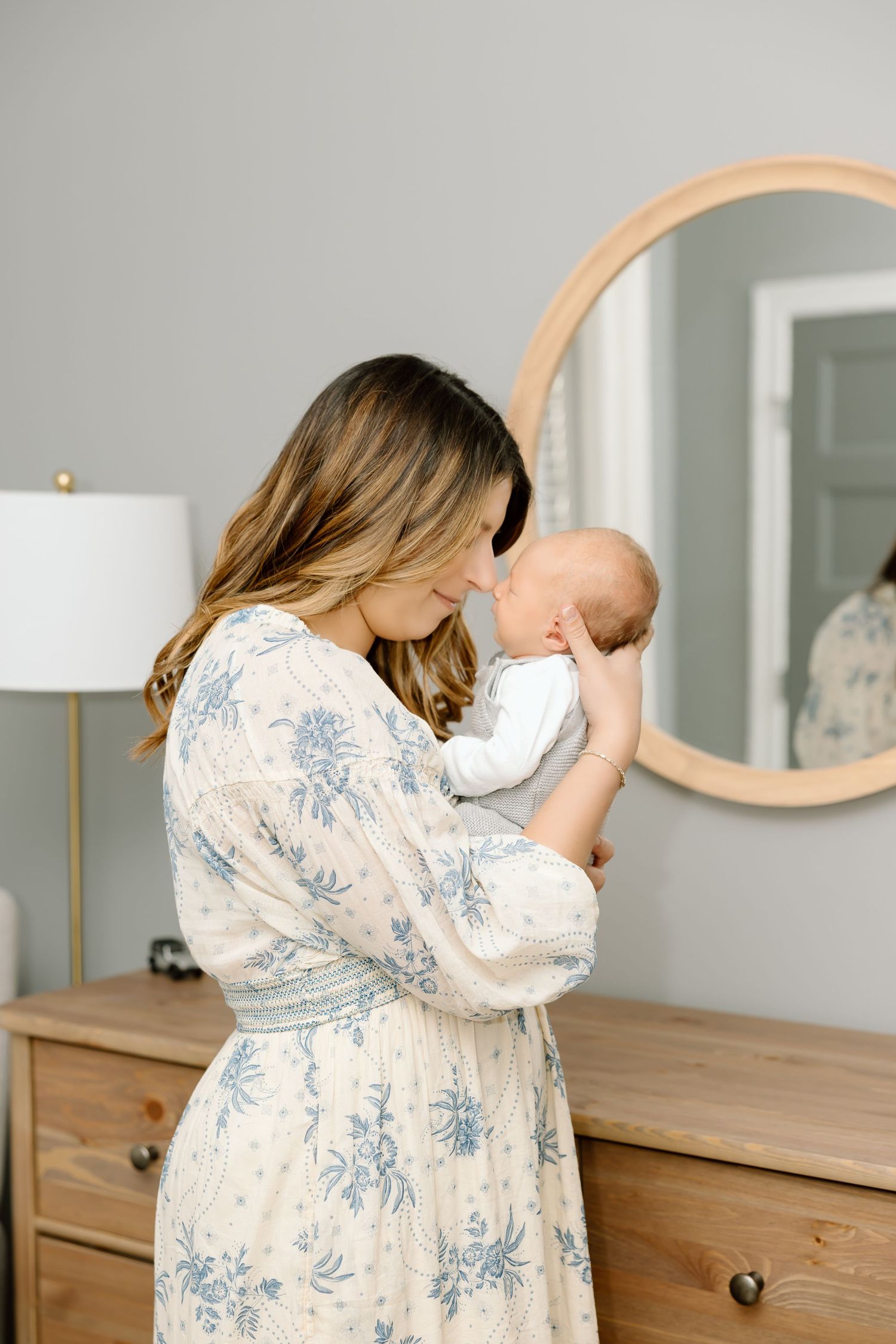 A parent in a floral dress holds and comforts a crying baby in a bright, modern bedroom setting.