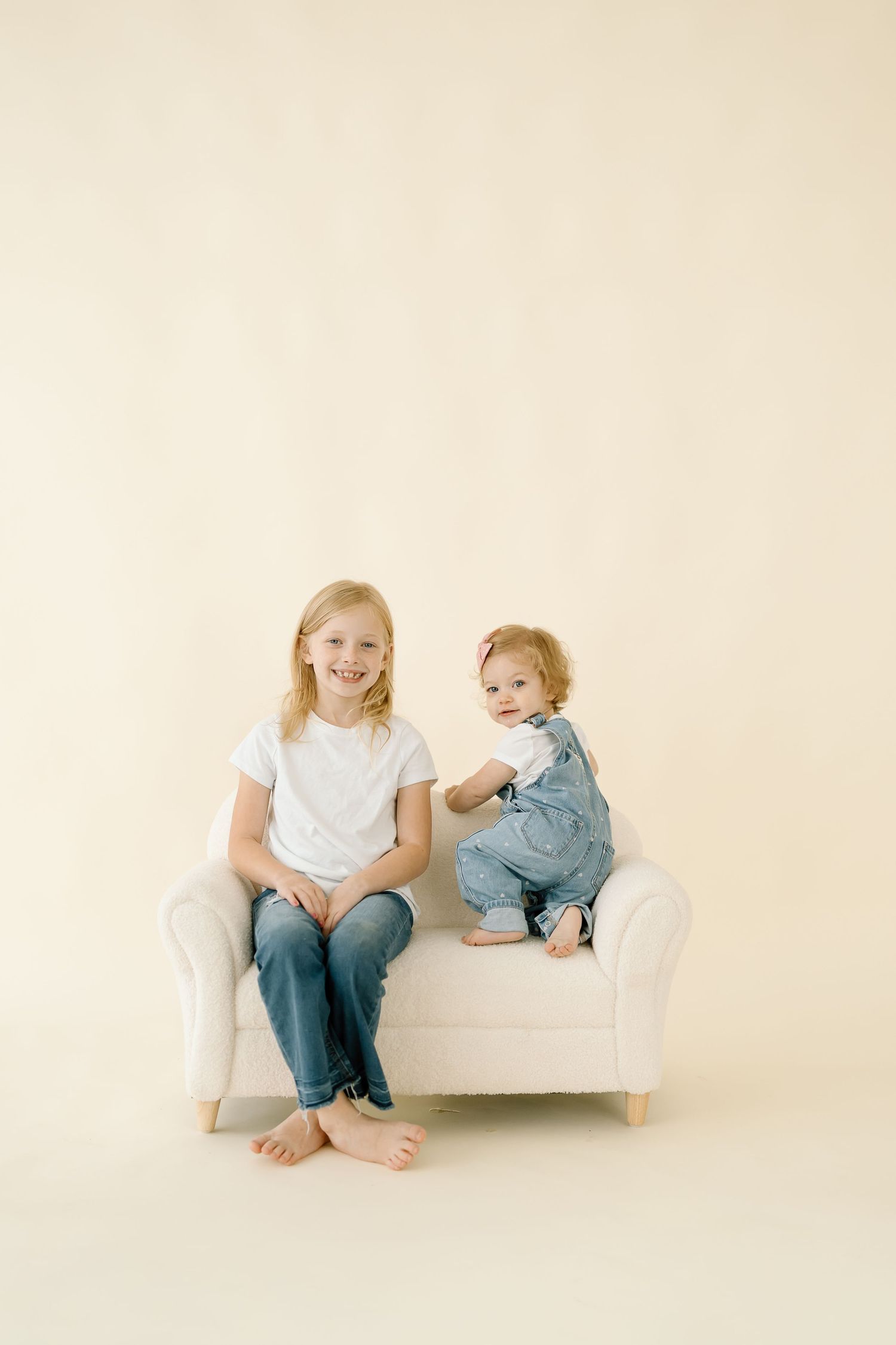 Two children sitting together on a white bench against a light neutral background in matching casual outfits.