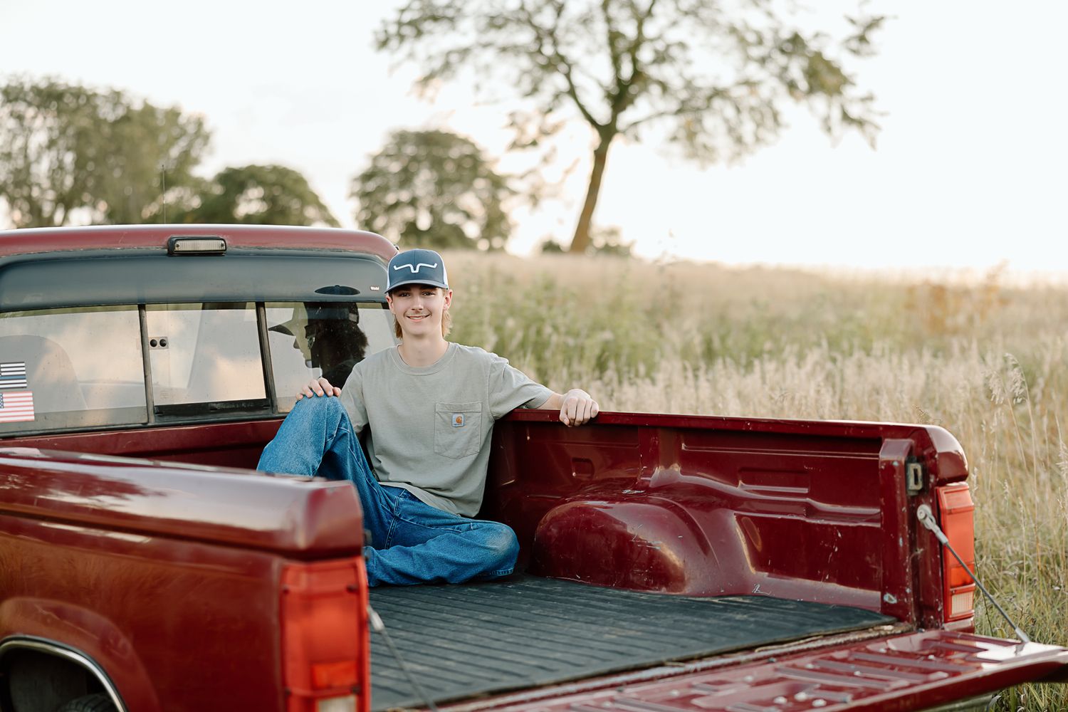 A vintage red pickup truck parked in a rural field at sunset with someone sitting in the truck bed.