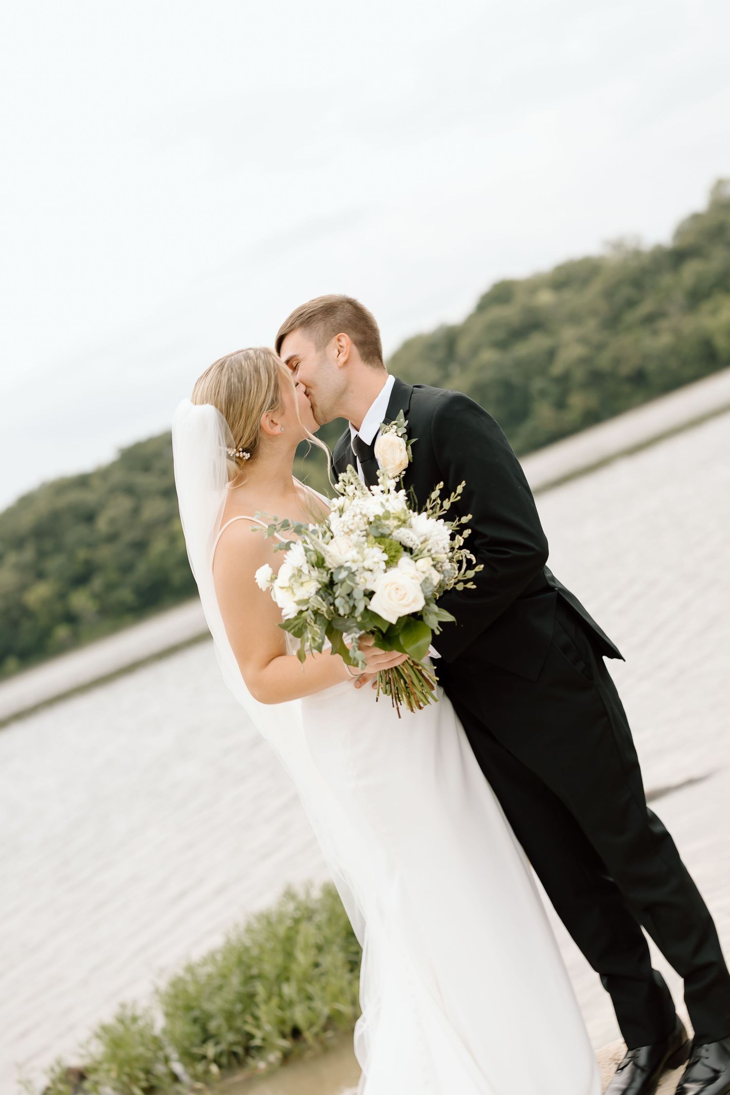 A romantic wedding couple shares a kiss while holding a white floral bouquet by the waterfront.