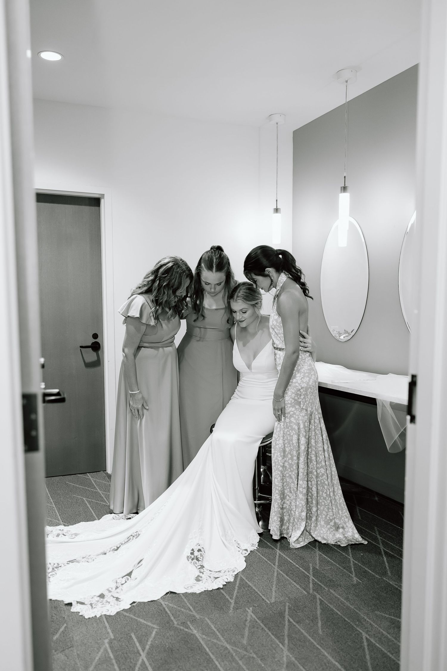 Wedding party in formal gowns gathered together in a modern dressing room with oval mirror and pendant lights.