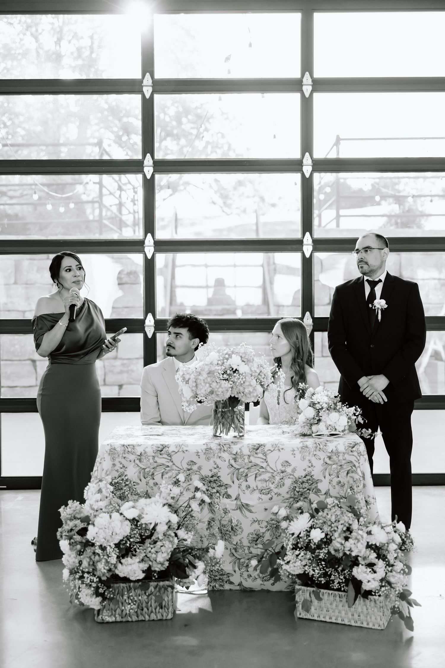 Black and white wedding reception photo with bride and groom at sweetheart table, bridesmaid giving toast, floral arrangements.
