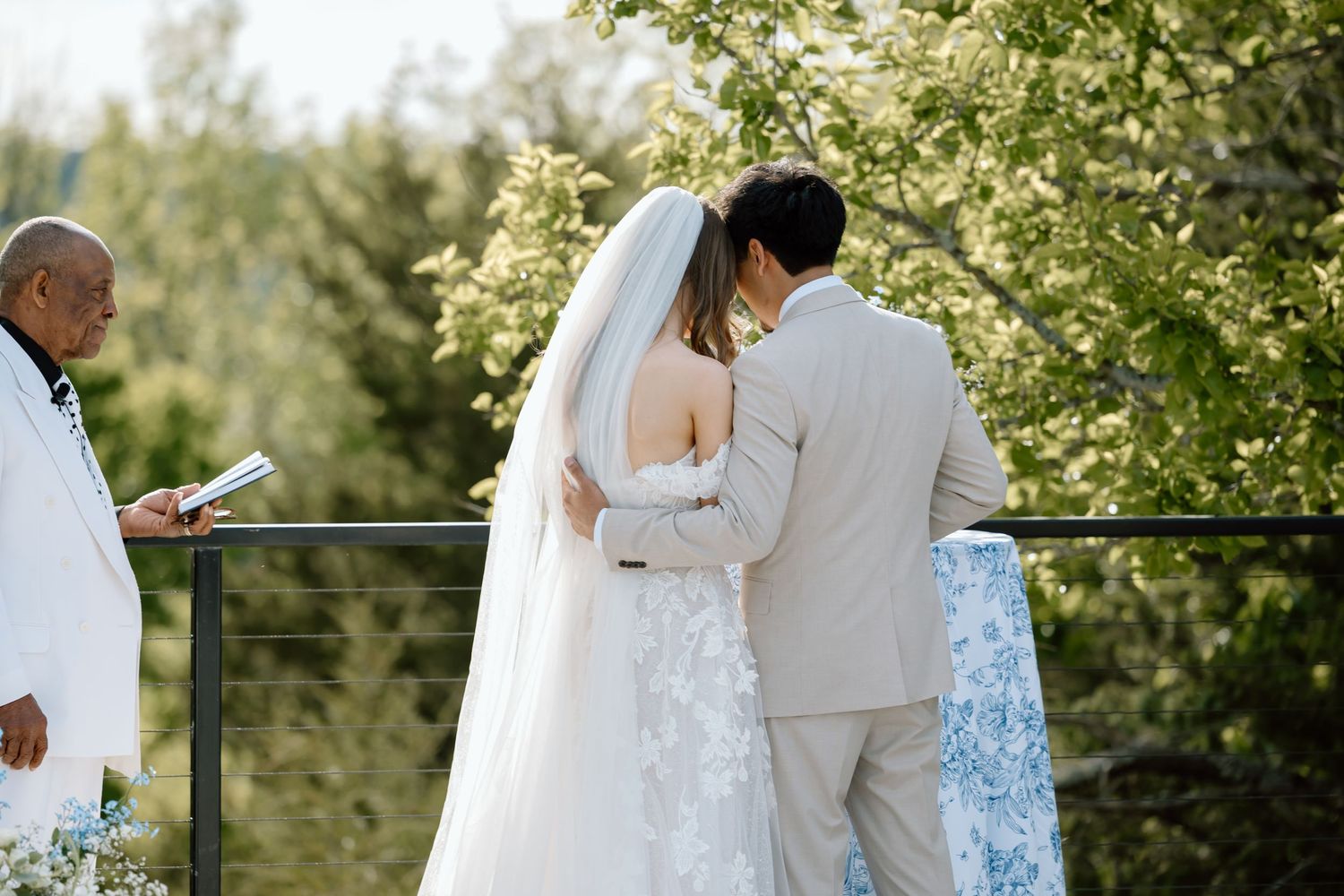 Bride and groom sharing a kiss during outdoor wedding ceremony with lush green trees in the background.