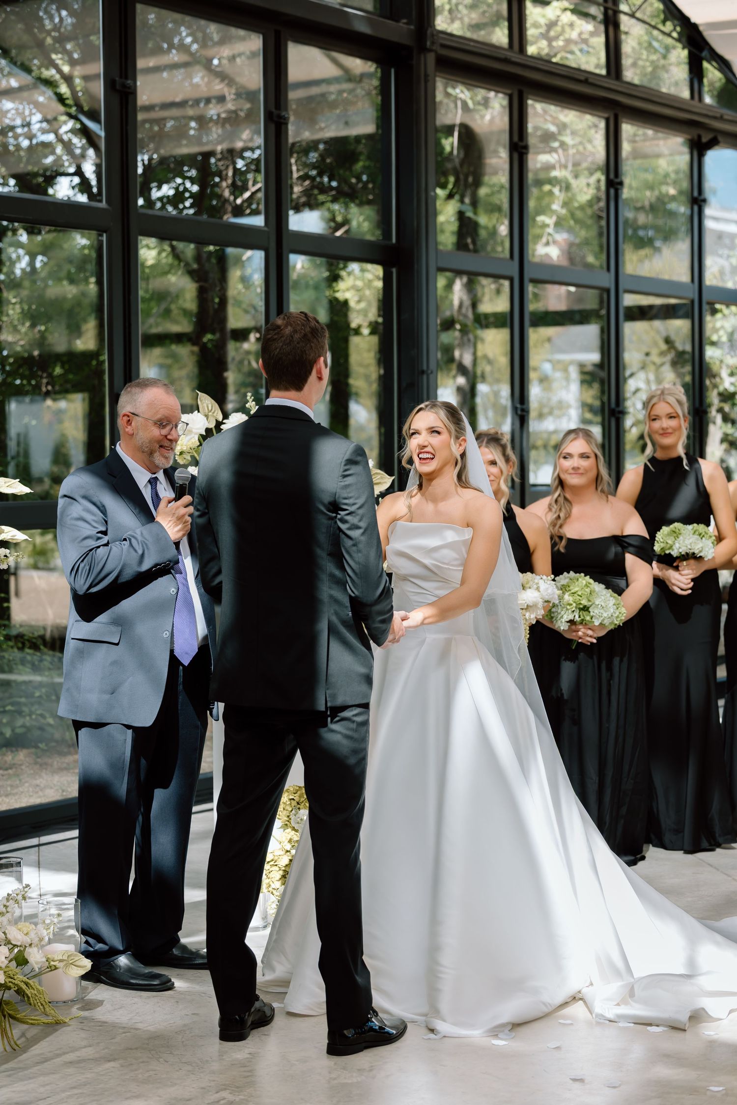 Bride and groom exchange vows at elegant indoor wedding ceremony with bridesmaids in black dresses and large glass windows.