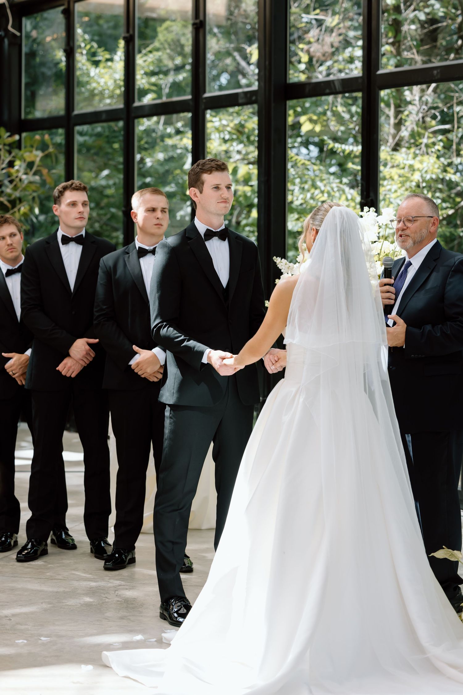 Bride and groom holding hands at wedding altar with groomsmen in black tuxedos and officiant in elegant venue.
