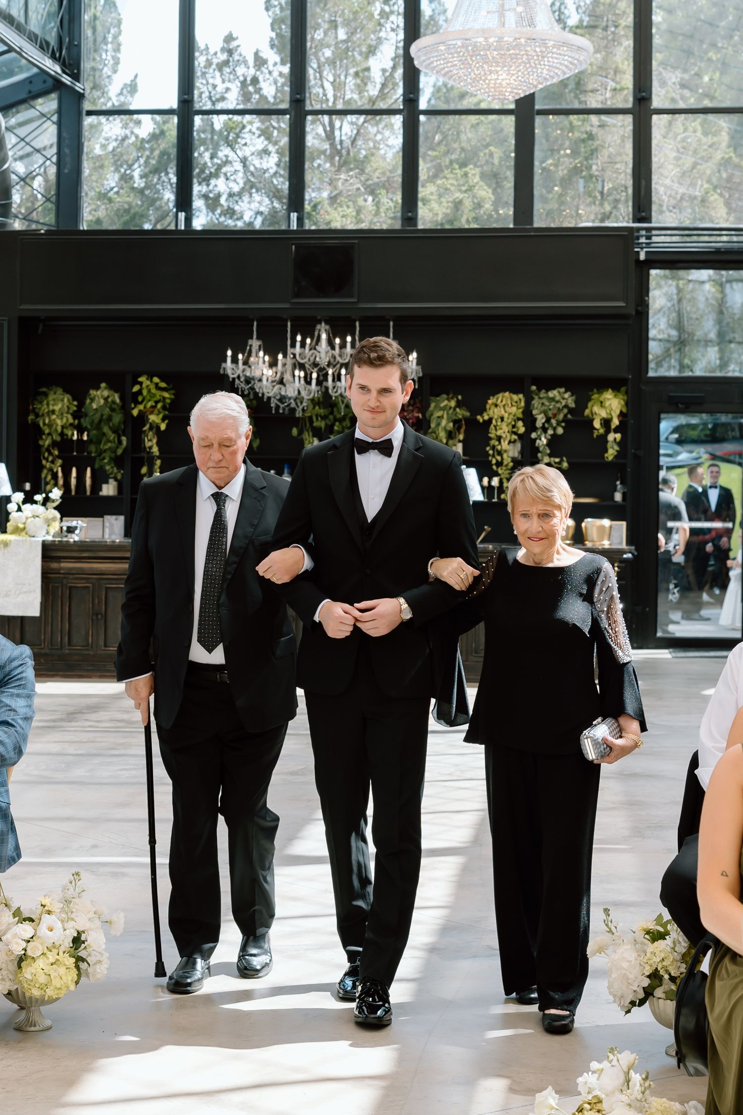 Groom in black tuxedo walks down wedding aisle escorted by his parents in elegant venue with chandelier.