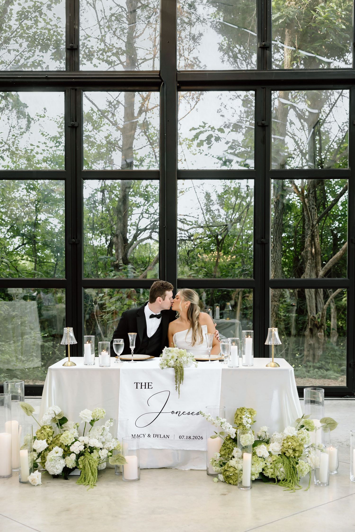 Bride and groom kissing at elegant sweetheart table with white florals and candles before large glass windows.