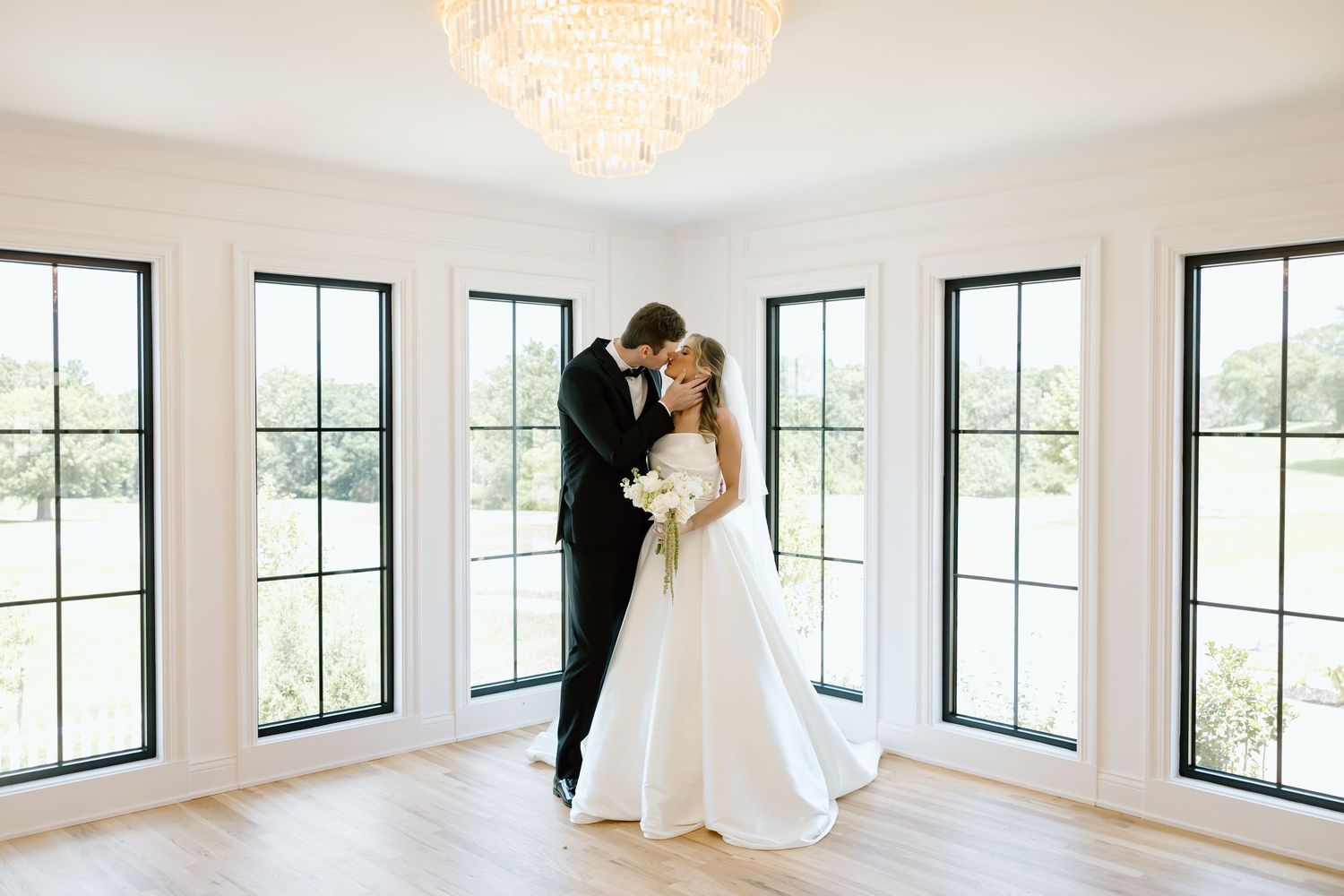 Bride and groom share a romantic kiss in a bright white room with large windows and elegant chandelier.