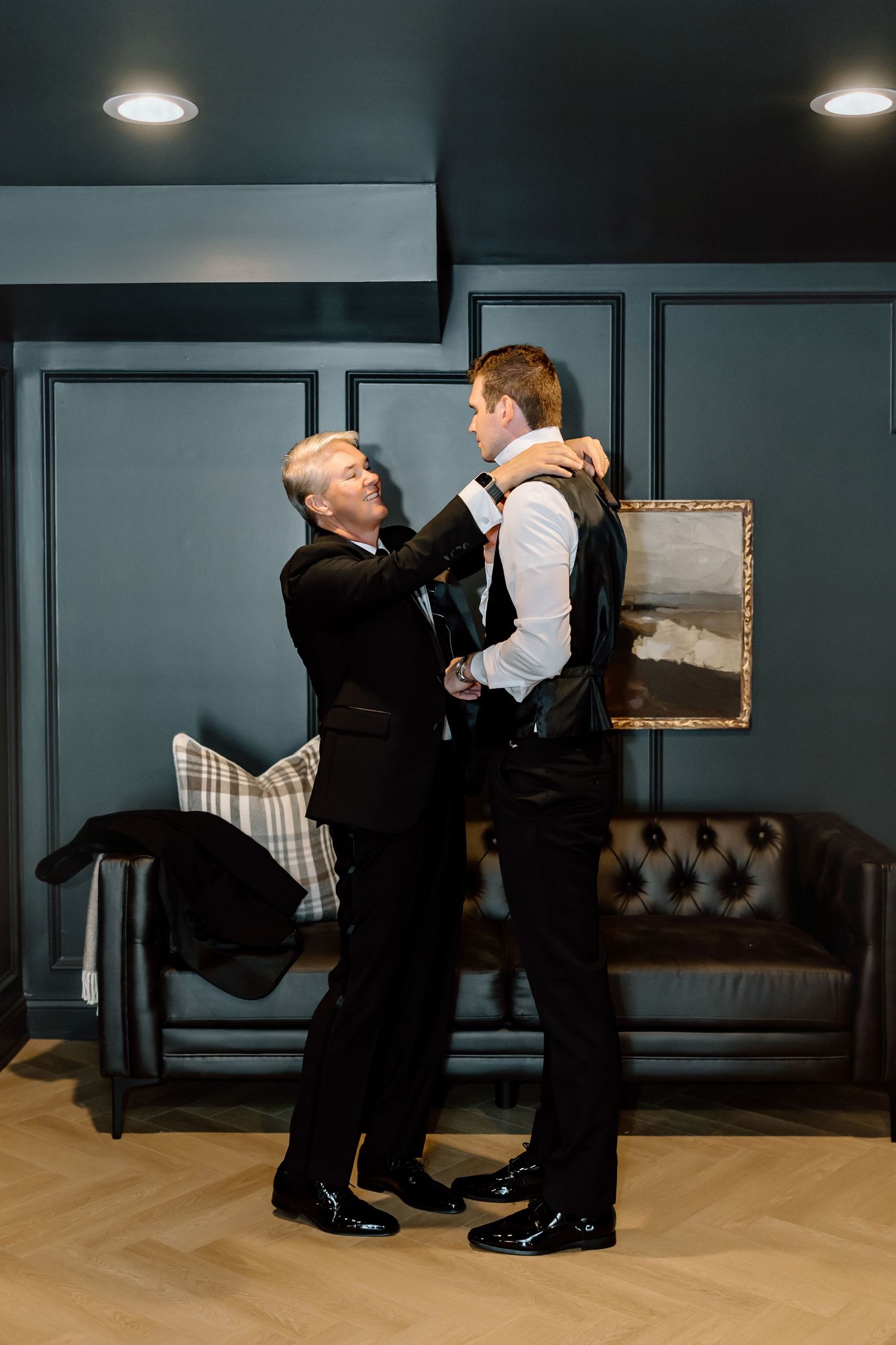 Two men in black suits helping each other get dressed in an elegant room before a formal event.