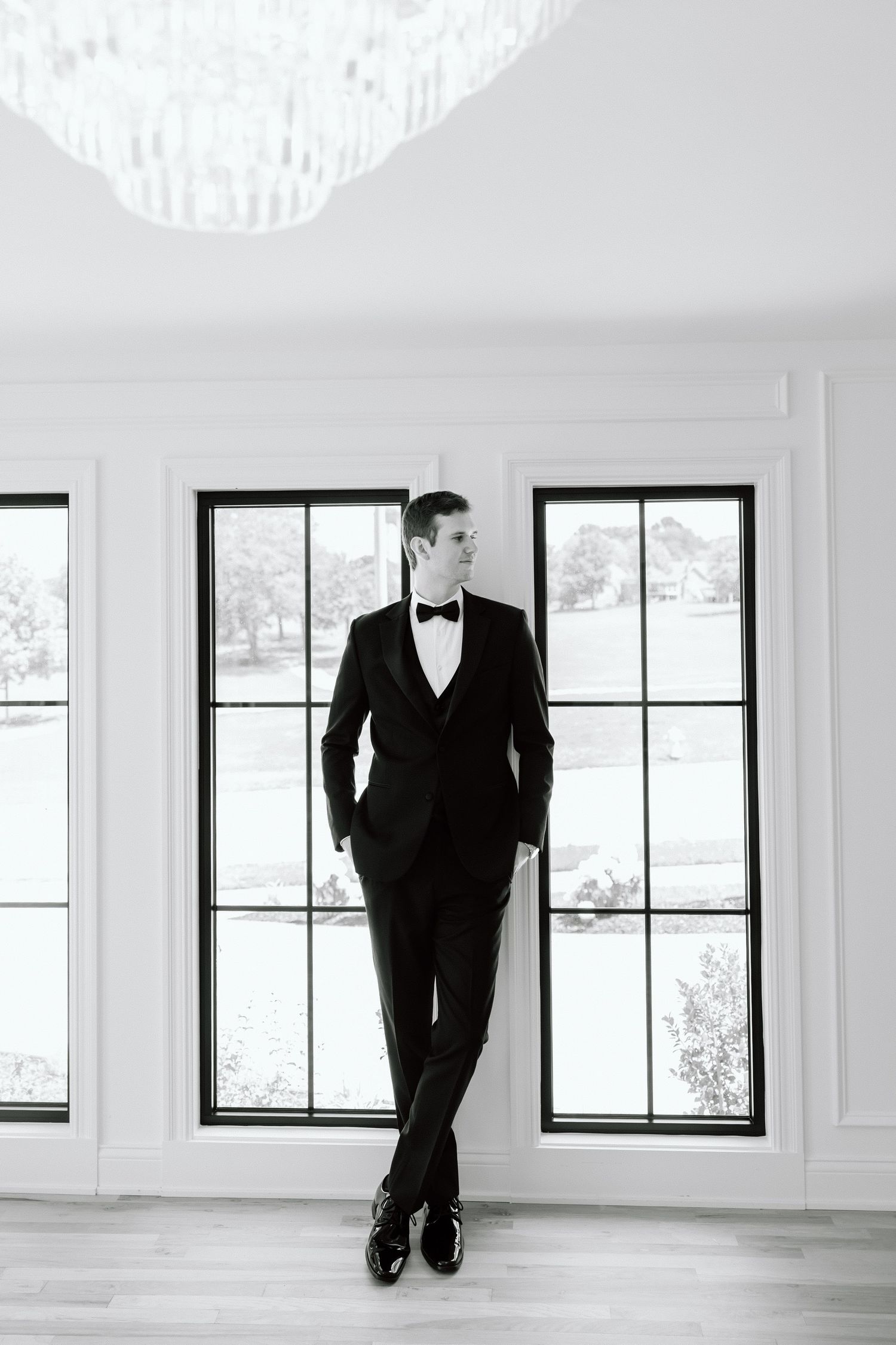 A groom in a black tuxedo and bow tie poses confidently between tall windows in an elegant white room.