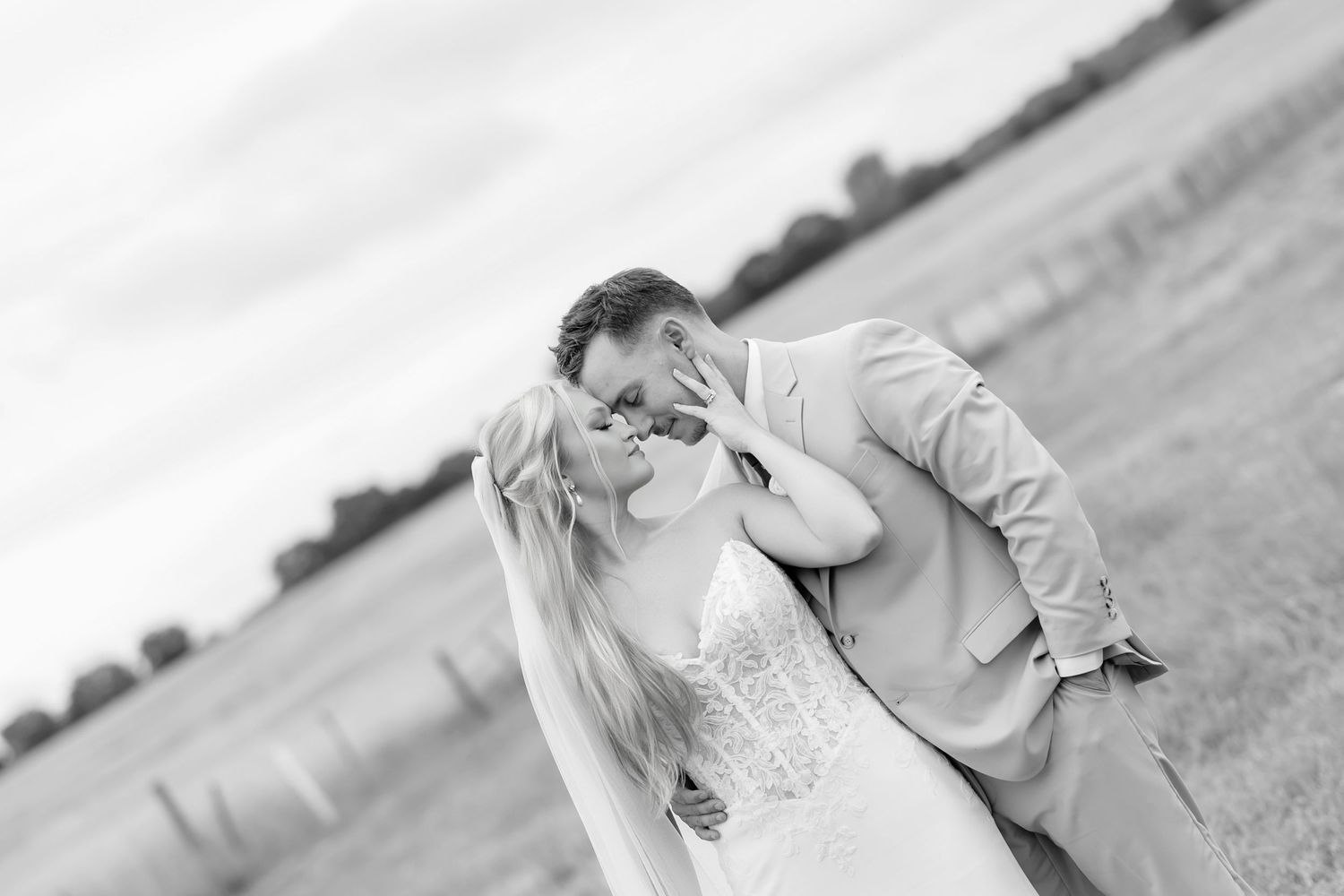 Black and white photo of a couple sharing a romantic kiss in an open field, bride in white gown and groom in suit.