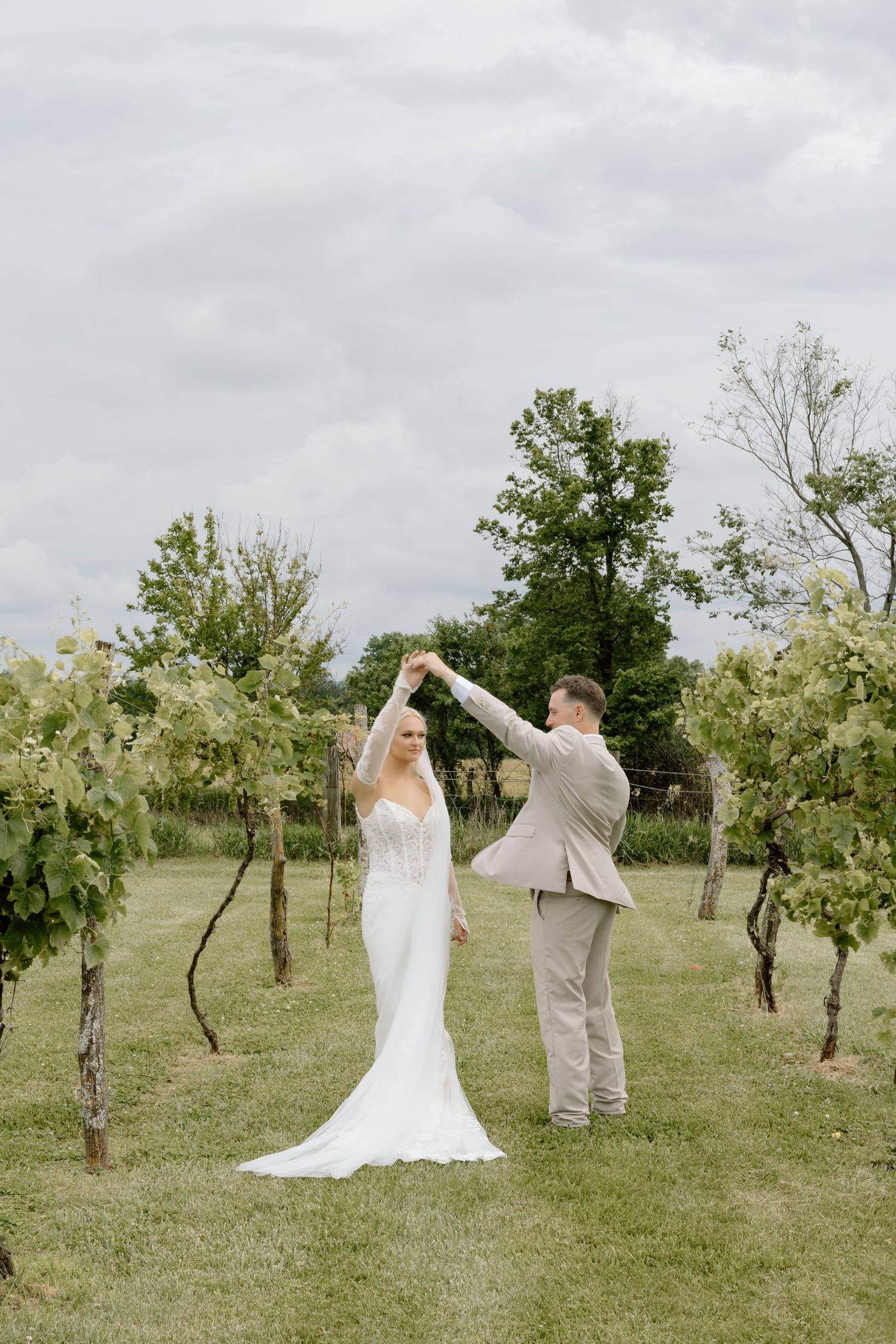 Bride and groom dancing in a lush vineyard, bride in a white gown with veil, groom in a beige suit.