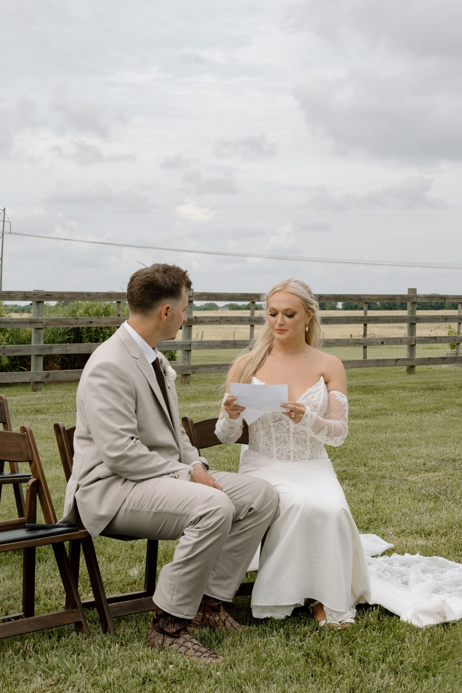 Bride in white lace gown reads vows to groom in tan suit during outdoor farm wedding ceremony.