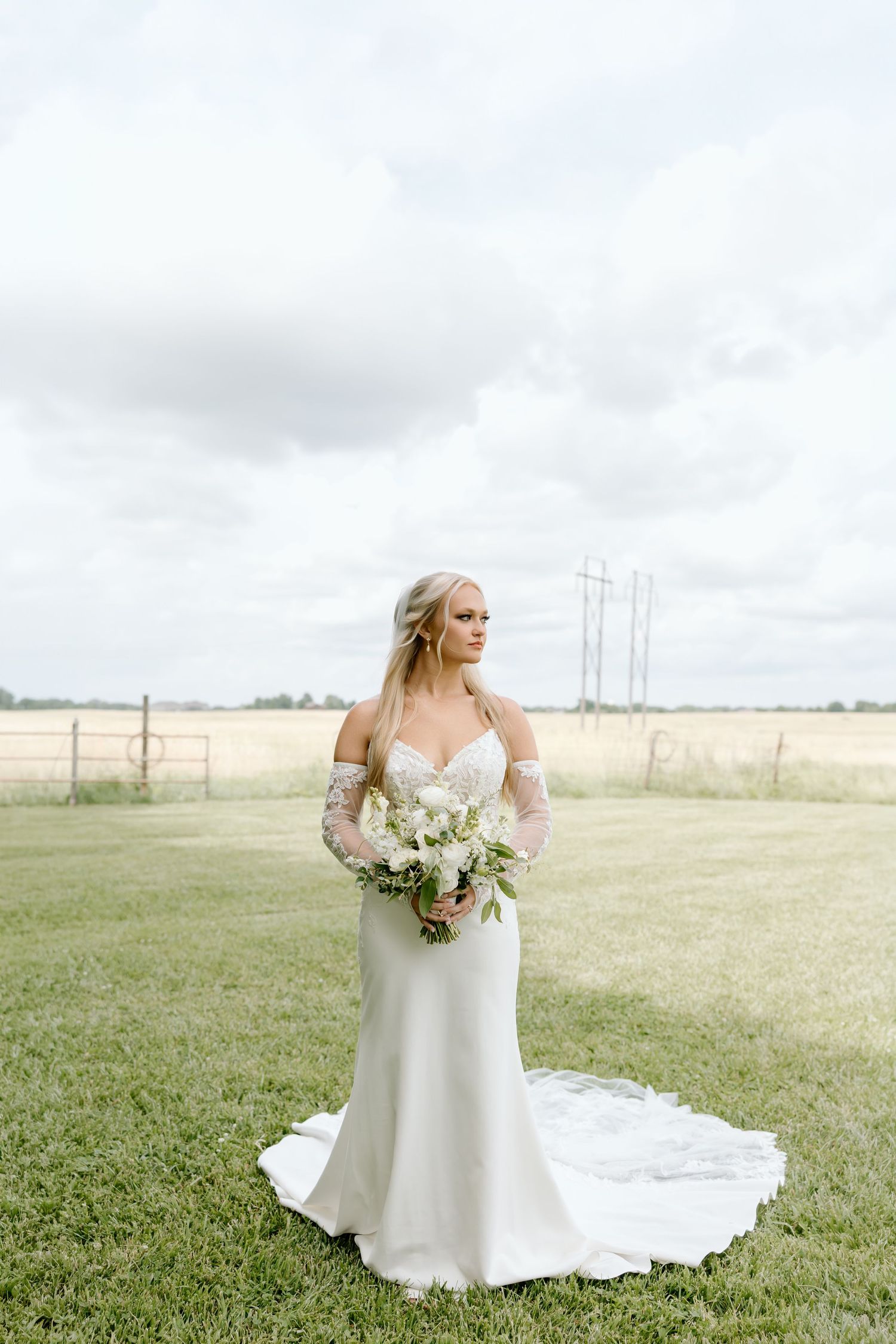 Bride in white fitted gown with lace sleeves holds floral bouquet in green field under cloudy sky.