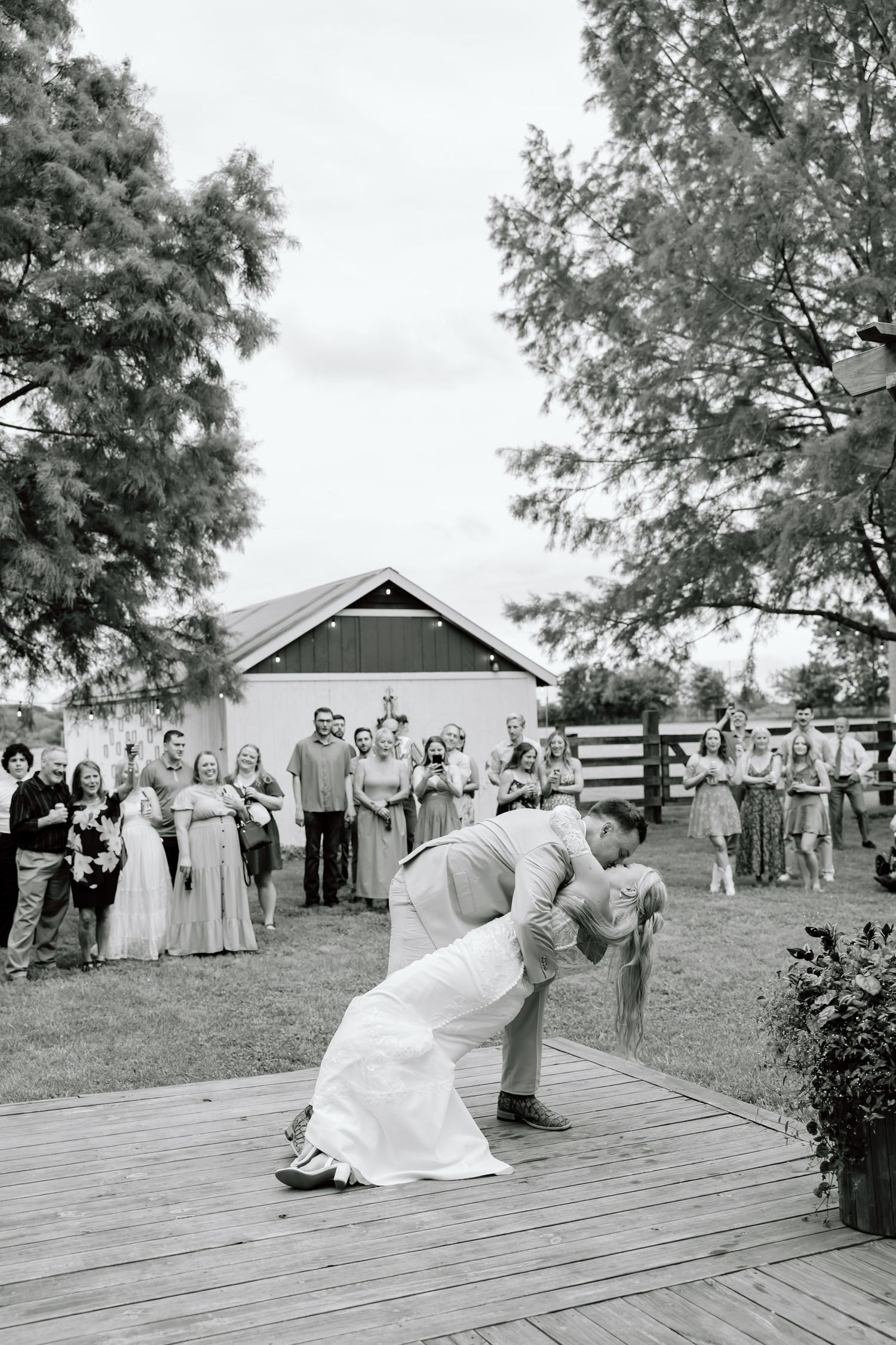 Black and white photo of groom dipping bride for a kiss on outdoor dance floor with wedding guests watching.
