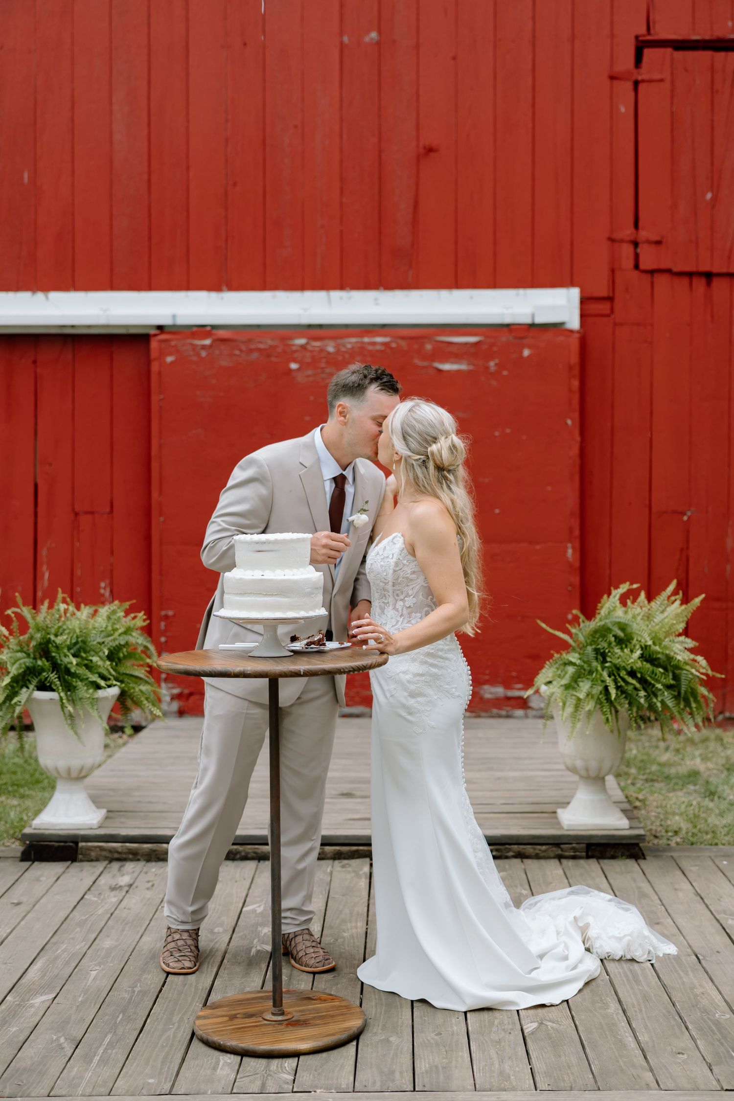 Bride and groom share a kiss beside their wedding cake in front of a rustic red barn on a wooden deck.