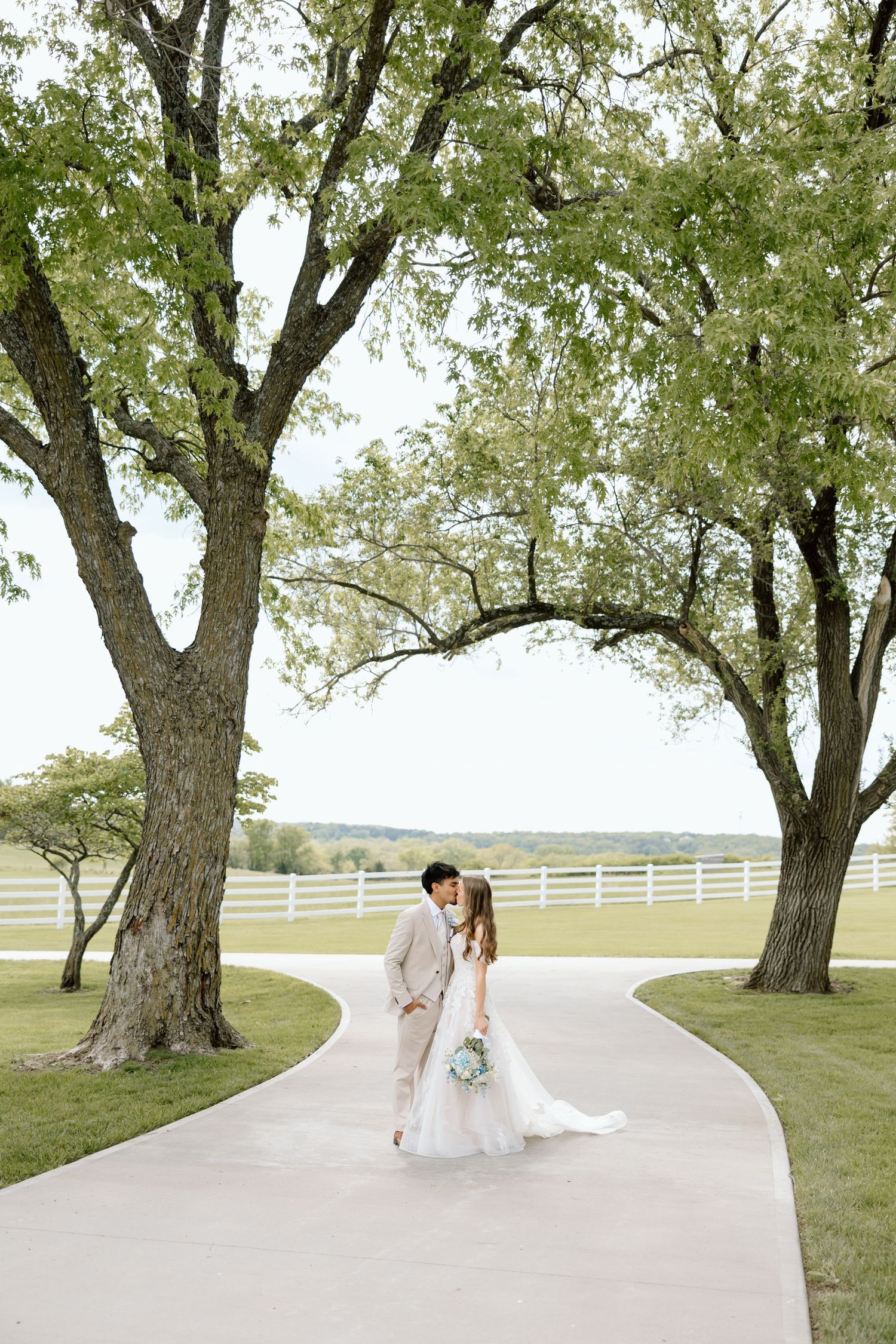 Bride and groom share a romantic kiss on a winding path flanked by large trees at an elegant outdoor wedding venue.