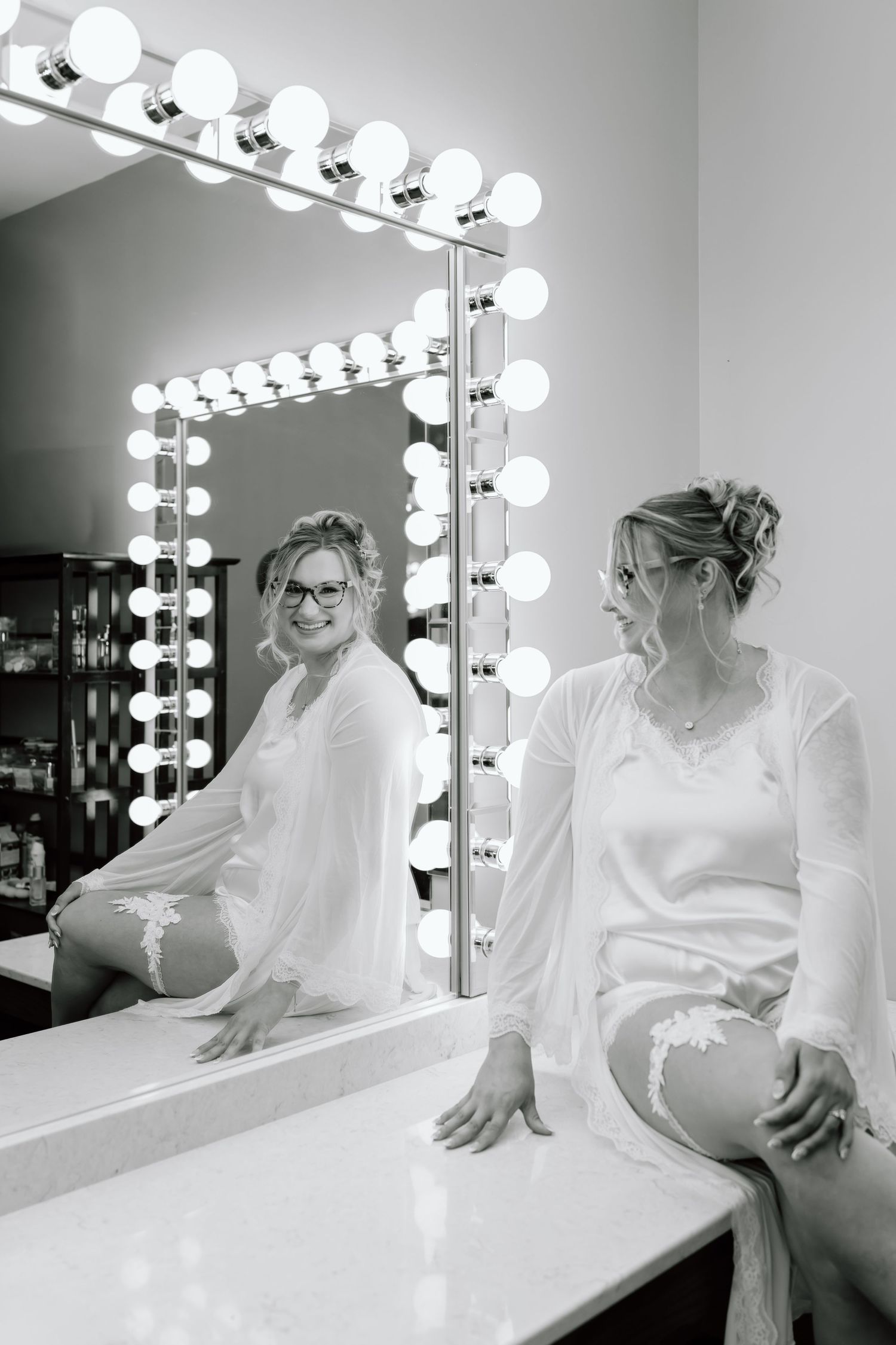 Black and white photo of someone in a white robe sitting at a vanity mirror with Hollywood-style lighting.