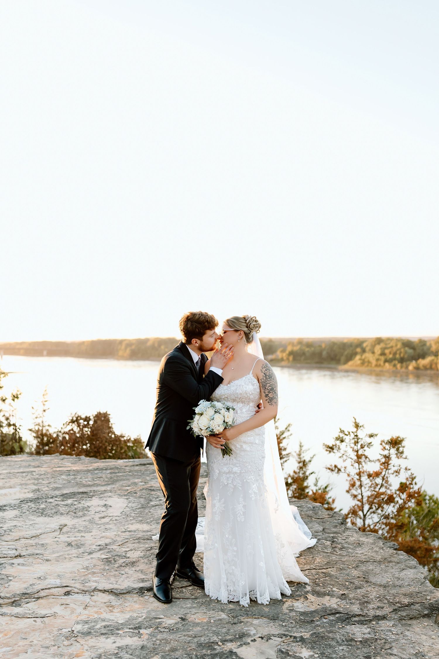 A romantic wedding couple embraces on a rocky cliff overlooking a serene lake at sunset.