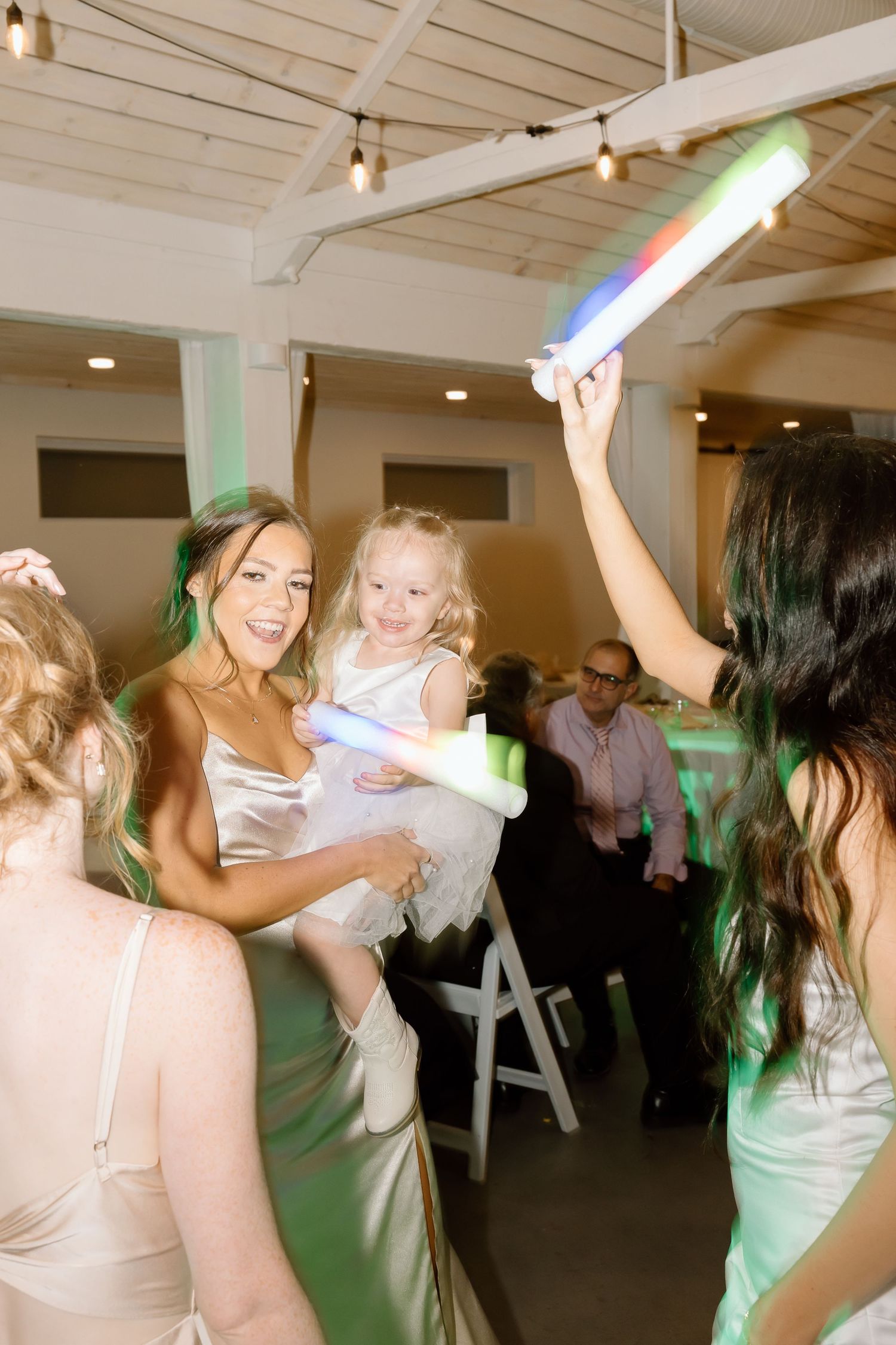 Party guests dancing and celebrating under string lights and colorful spotlights in an indoor venue.