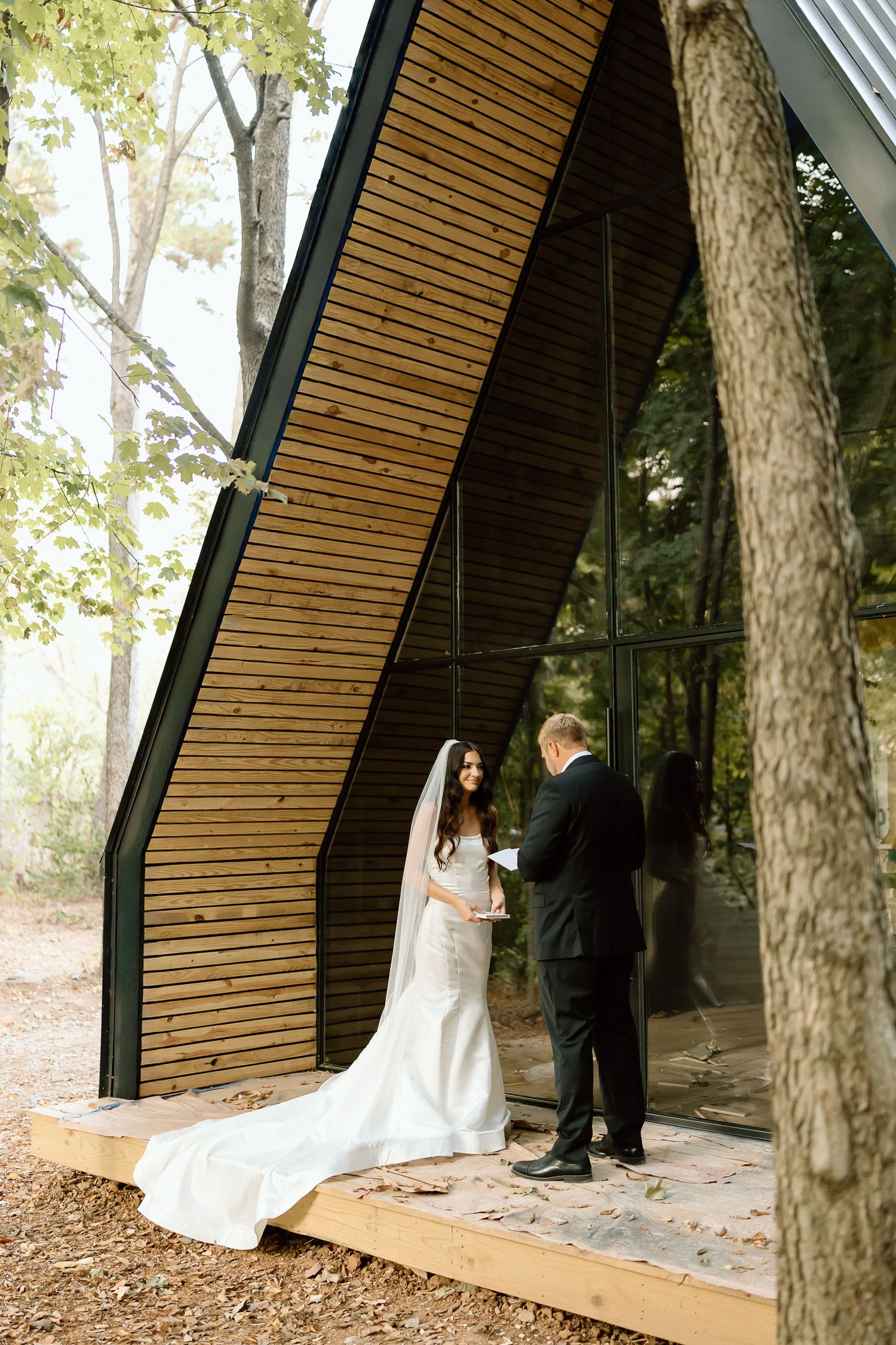 A wedding ceremony takes place in a modern wooden chapel structure nestled among tall trees in a forest setting.
