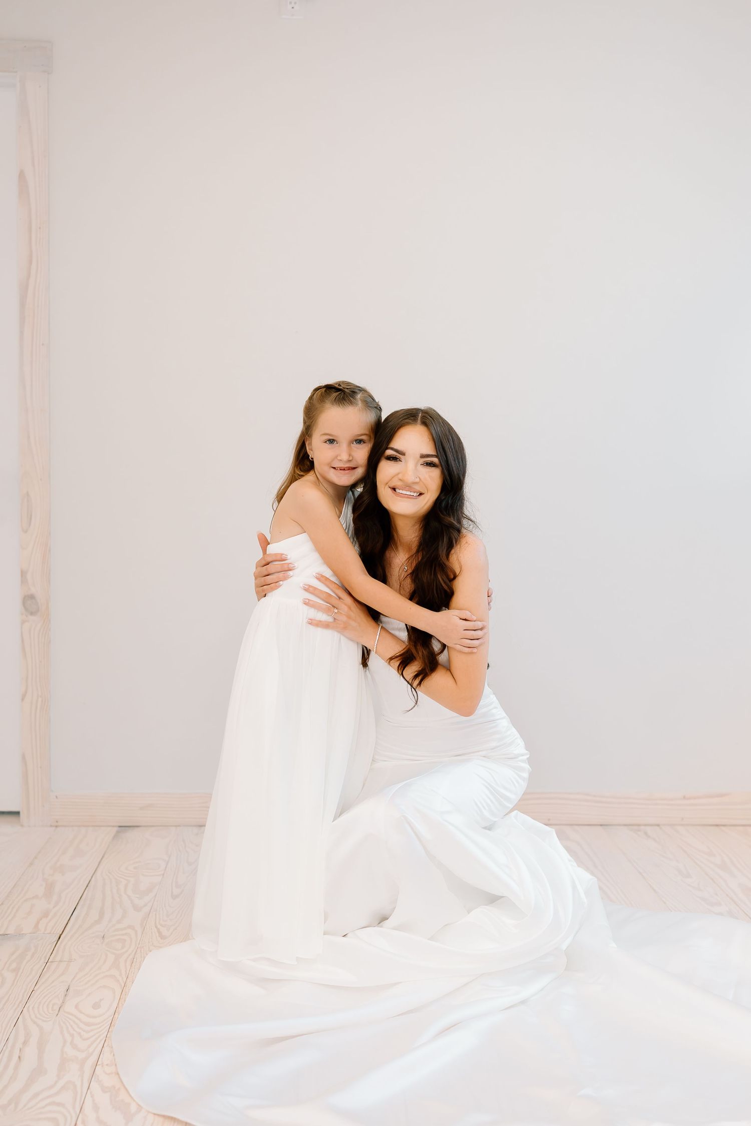 Two people in white dresses embrace while sitting on a light wooden floor in a bright minimalist studio setting.