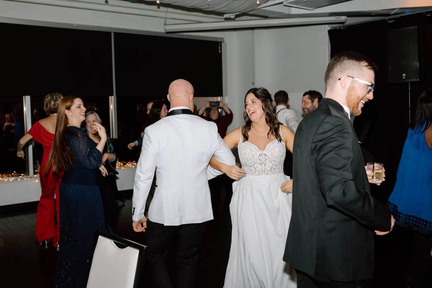 Wedding guests dancing and celebrating at an evening reception with a bride in a white dress on the dance floor.