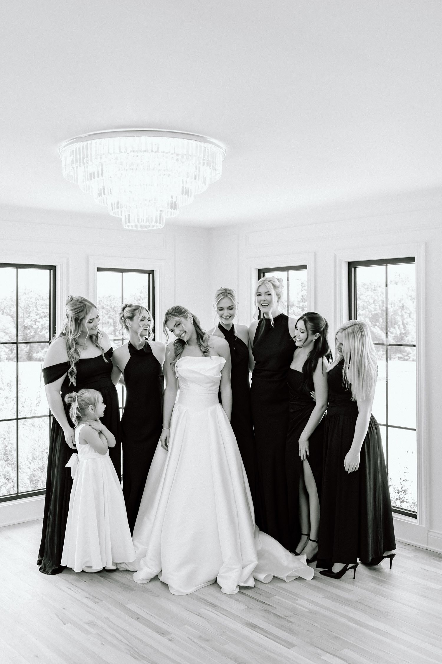 Black and white photo of a bride in a white gown surrounded by bridesmaids in black dresses and a flower girl in a bright room.
