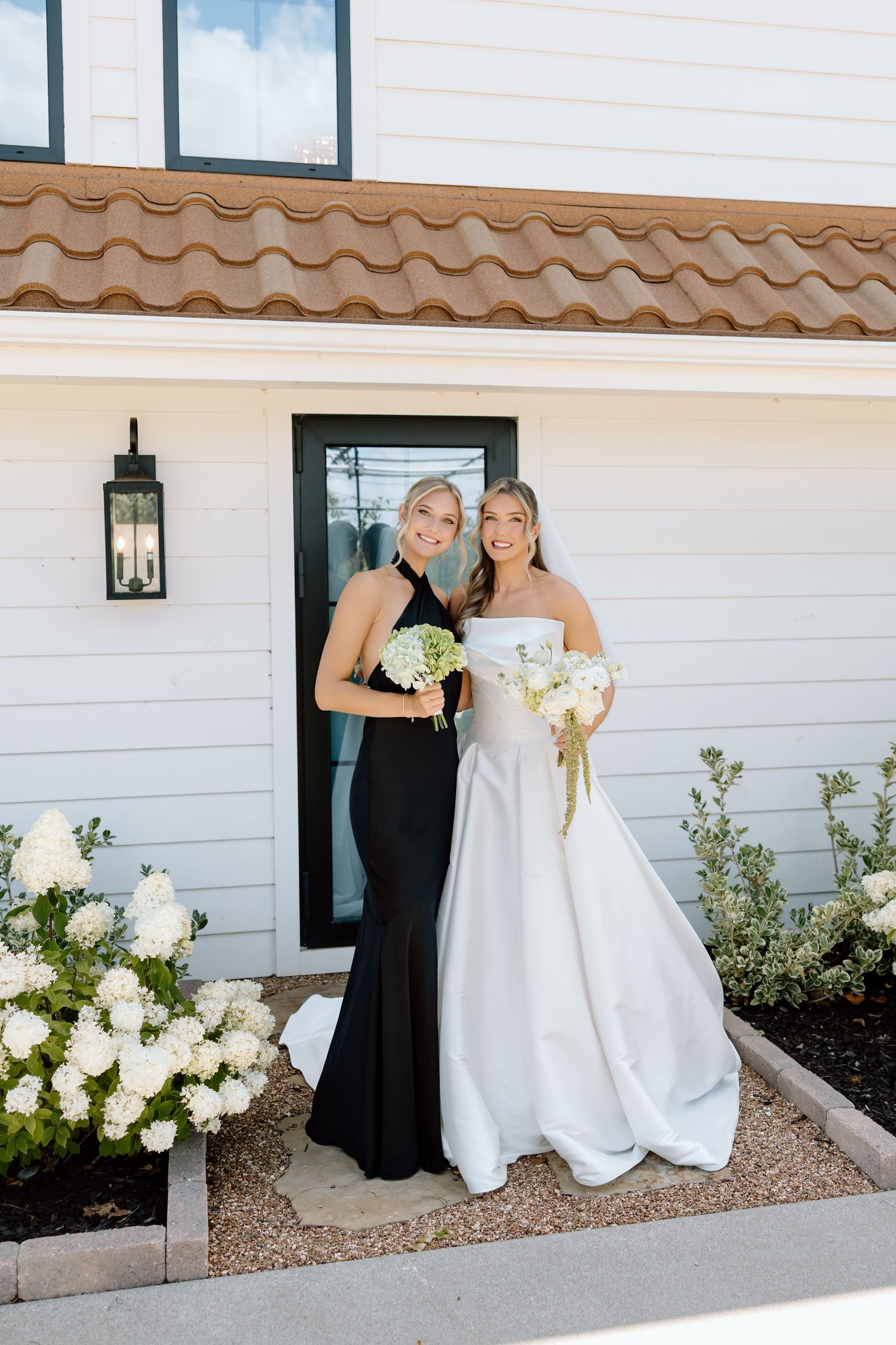 Bride in white off-shoulder gown and bridesmaid in black halter dress holding white bouquets outside a white home.