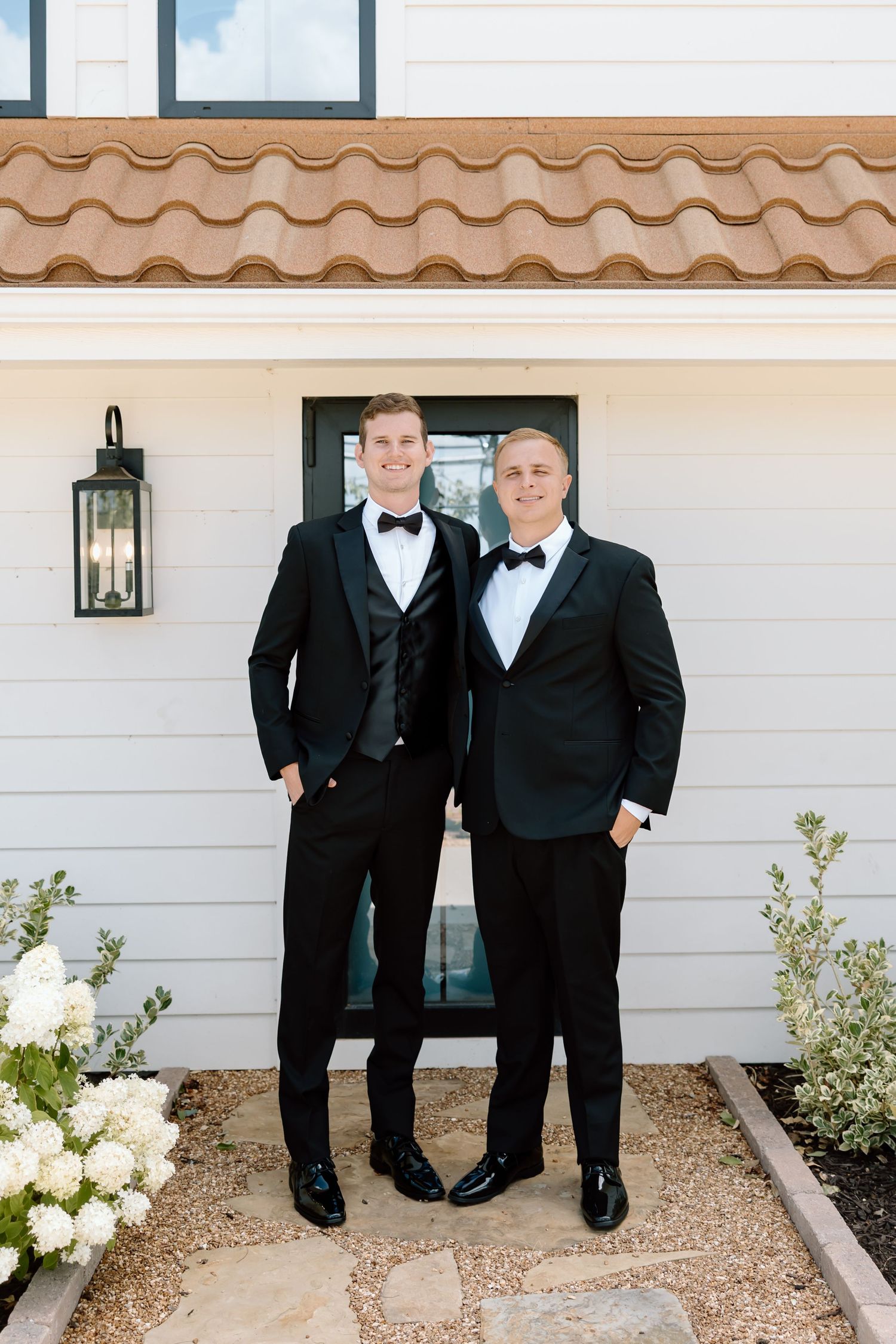 Two men in black tuxedos with bow ties pose together outside a white home with terra cotta roof tiles.
