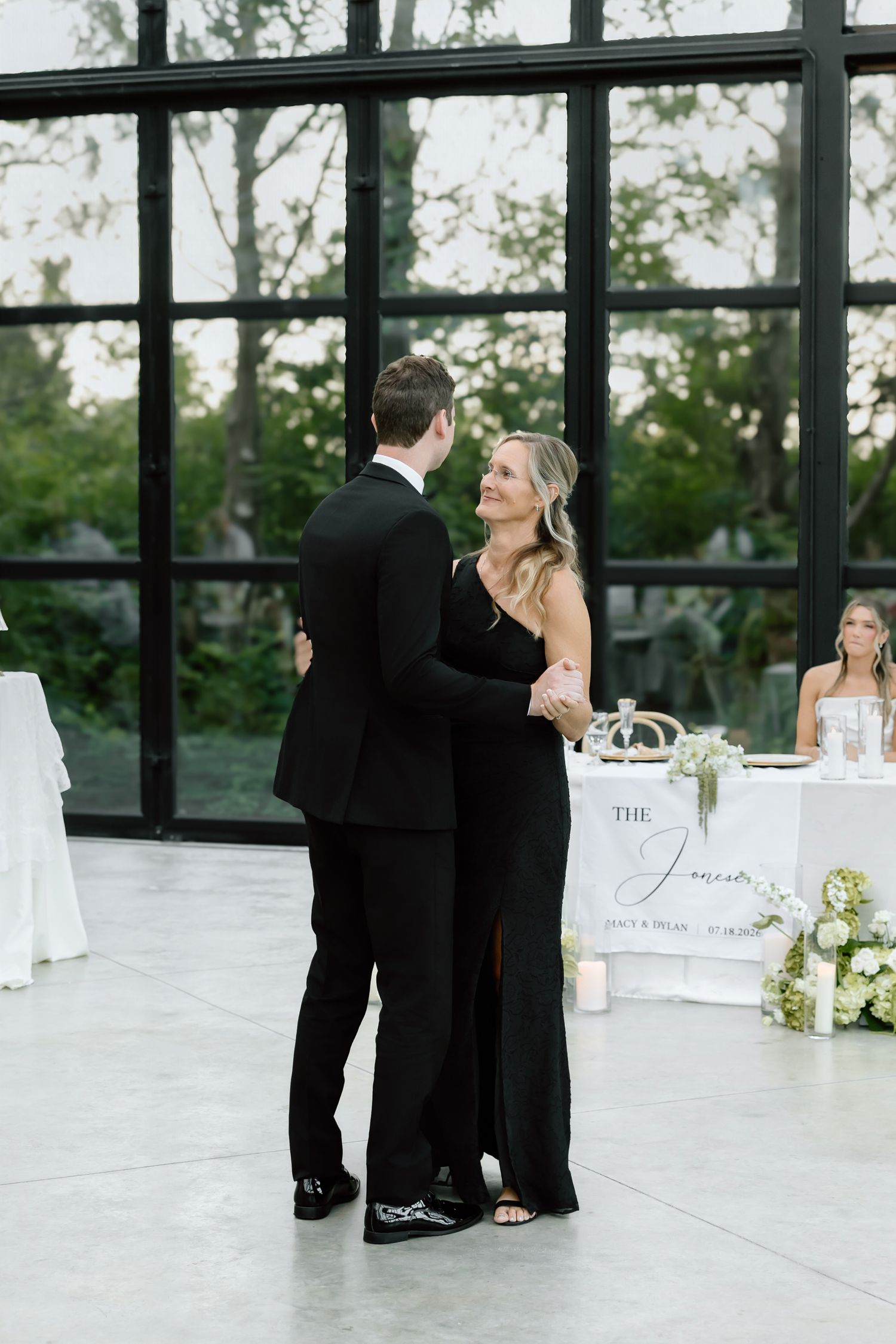 Couple sharing first dance at wedding reception in black formal attire, with elegant white floral decor and large windows behind them.