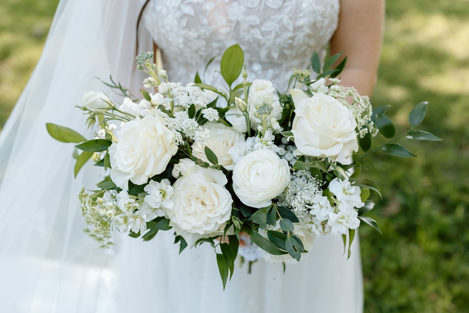 A classic white bridal bouquet with roses, baby's breath and green foliage held against a white wedding dress.