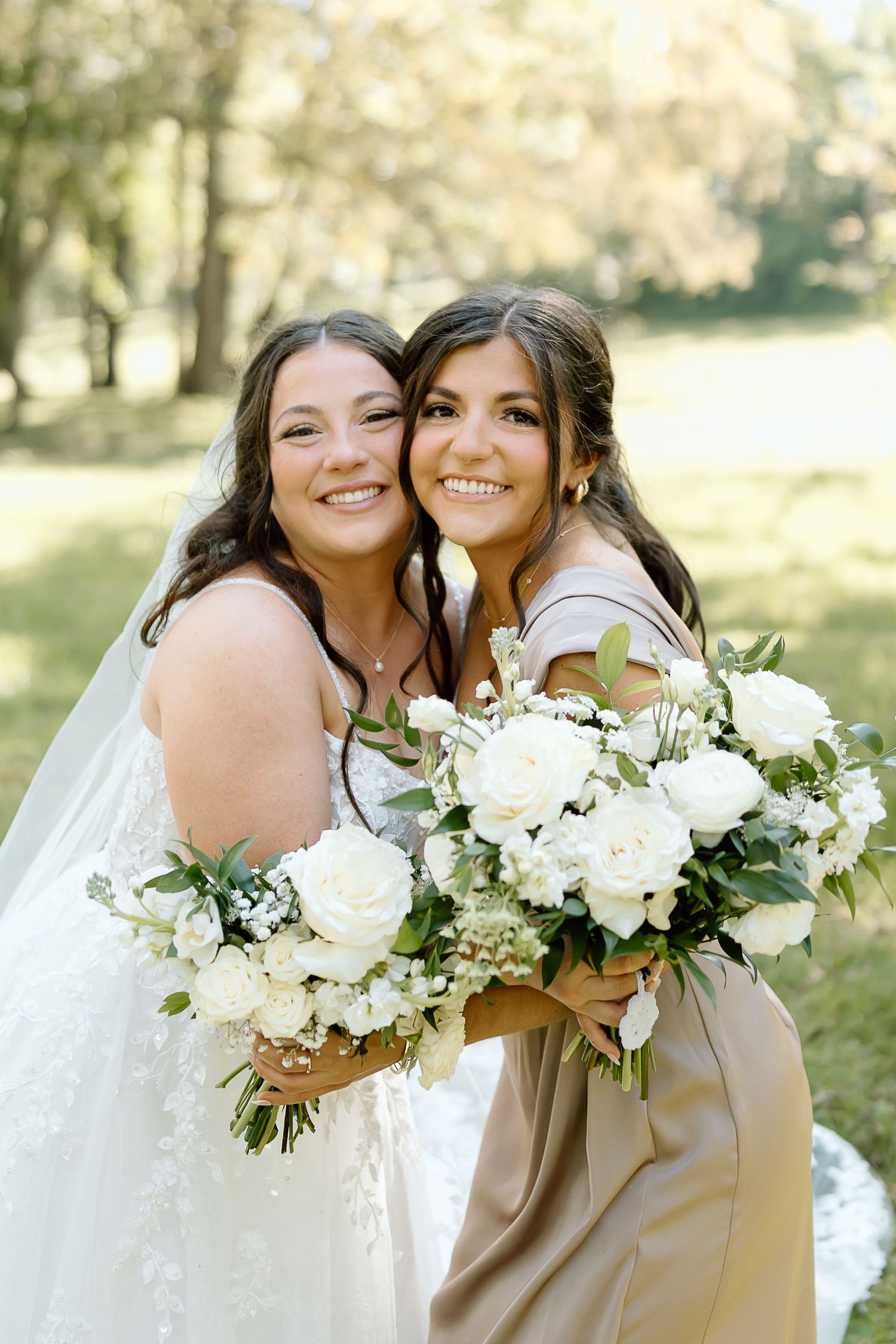 Bridesmaids in beige dresses holding elegant white floral bouquets pose together outdoors on a sunny day.