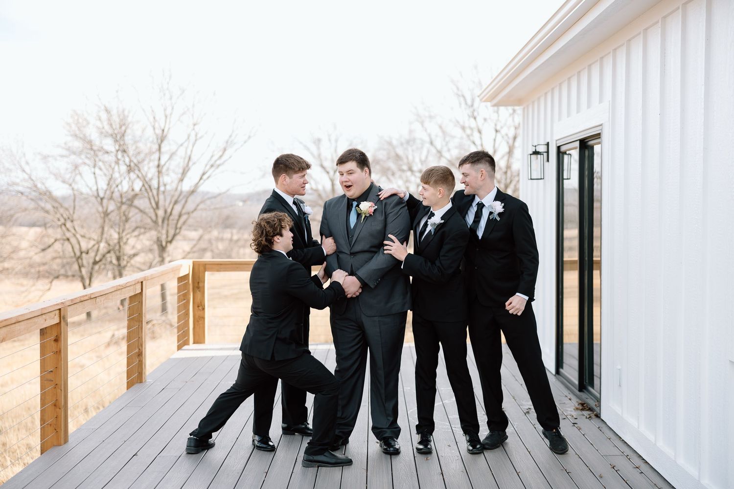 Wedding party in black suits stands together laughing and sharing a joyful moment outdoors.