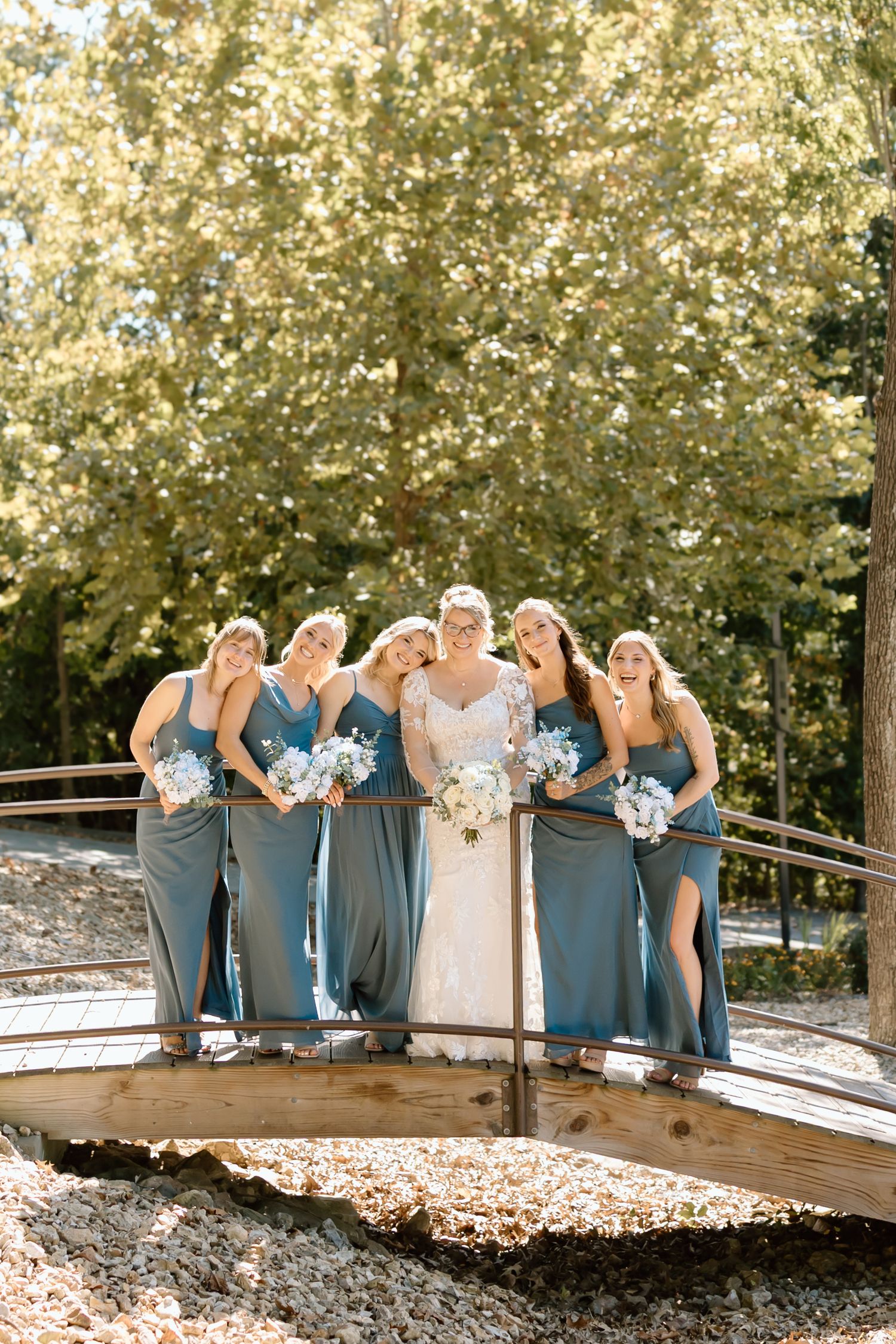 Wedding party in slate blue dresses pose together on wooden deck surrounded by autumn foliage.