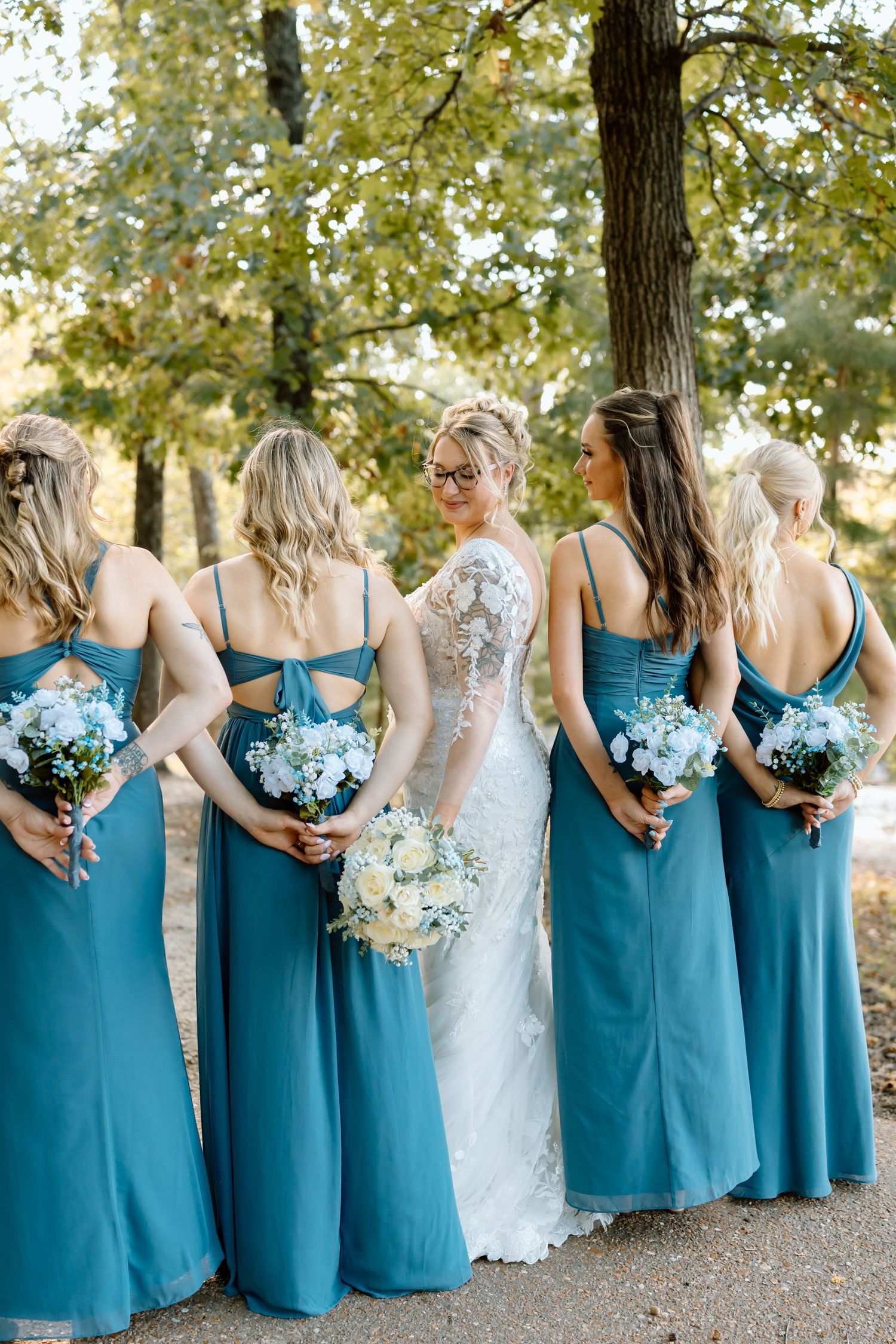 Bridesmaids in matching teal dresses stand in a row holding white and blue bouquets beside the bride in white lace.