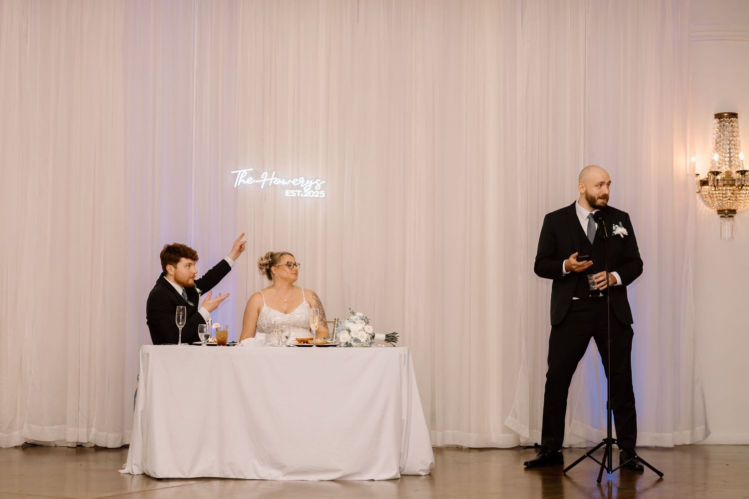 Wedding party members dance together during a reception in an elegant white-draped ballroom.