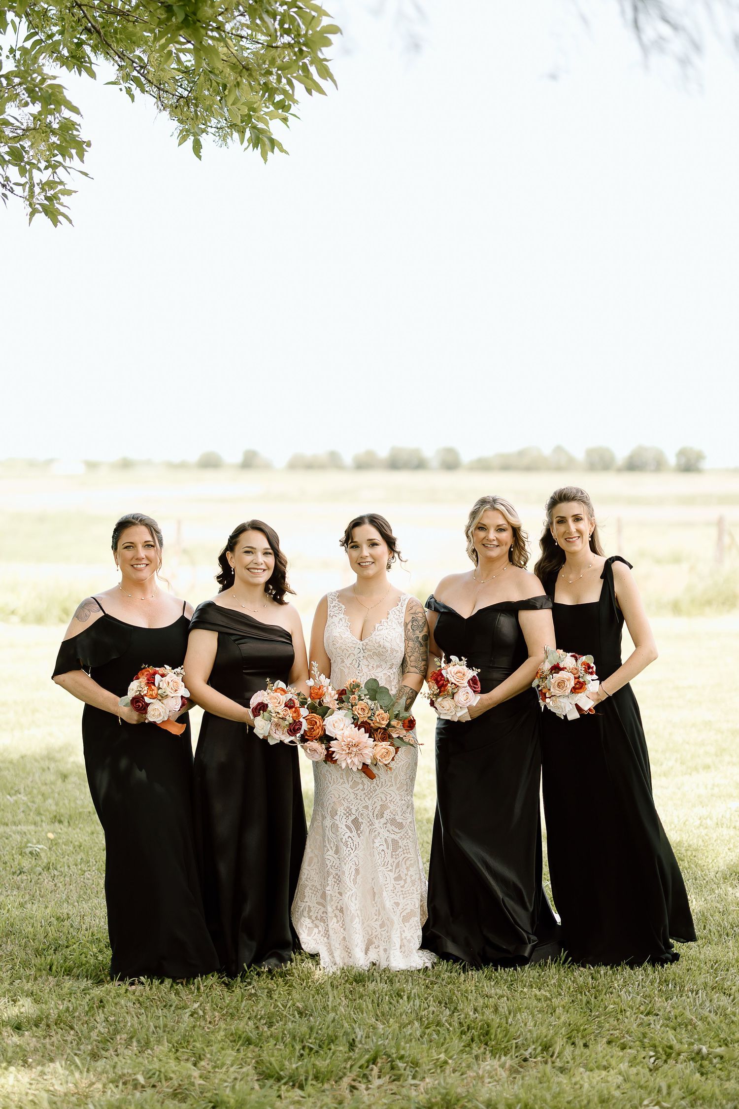 Wedding party in black dresses and white gown pose together outdoors on a grassy field with trees in background.