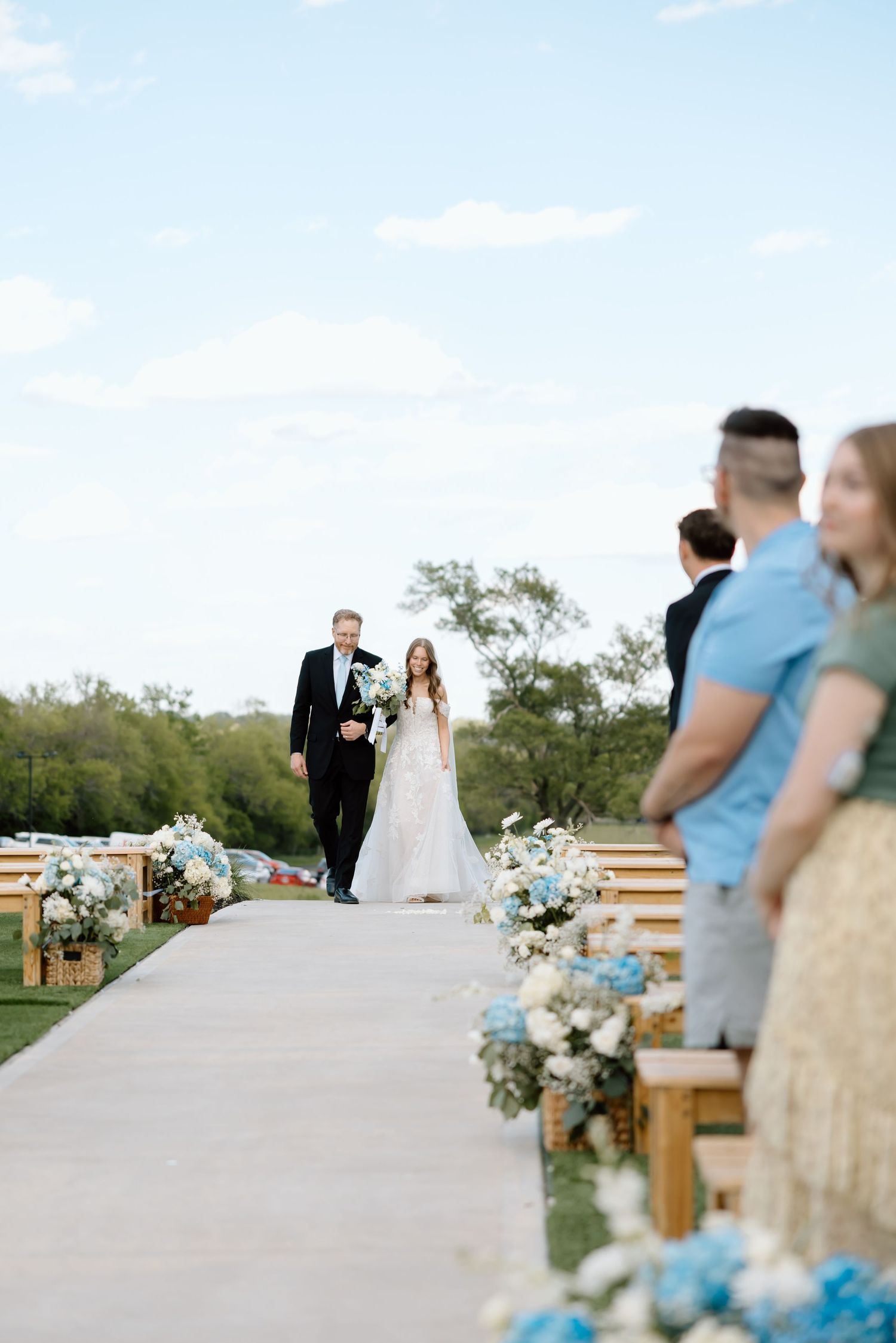 Bride walks down outdoor aisle with escort, flanked by blue and white floral arrangements and wooden benches with guests standing.