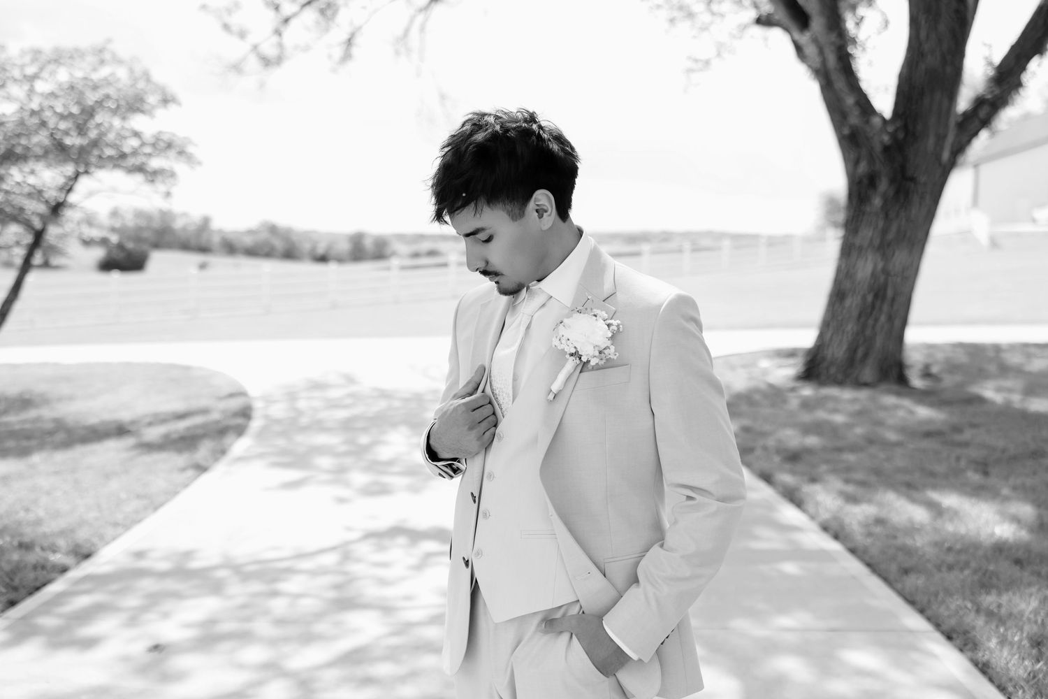 Groom in a light beige suit adjusting his cufflinks outdoors, with a white boutonniere, in a black and white photo.