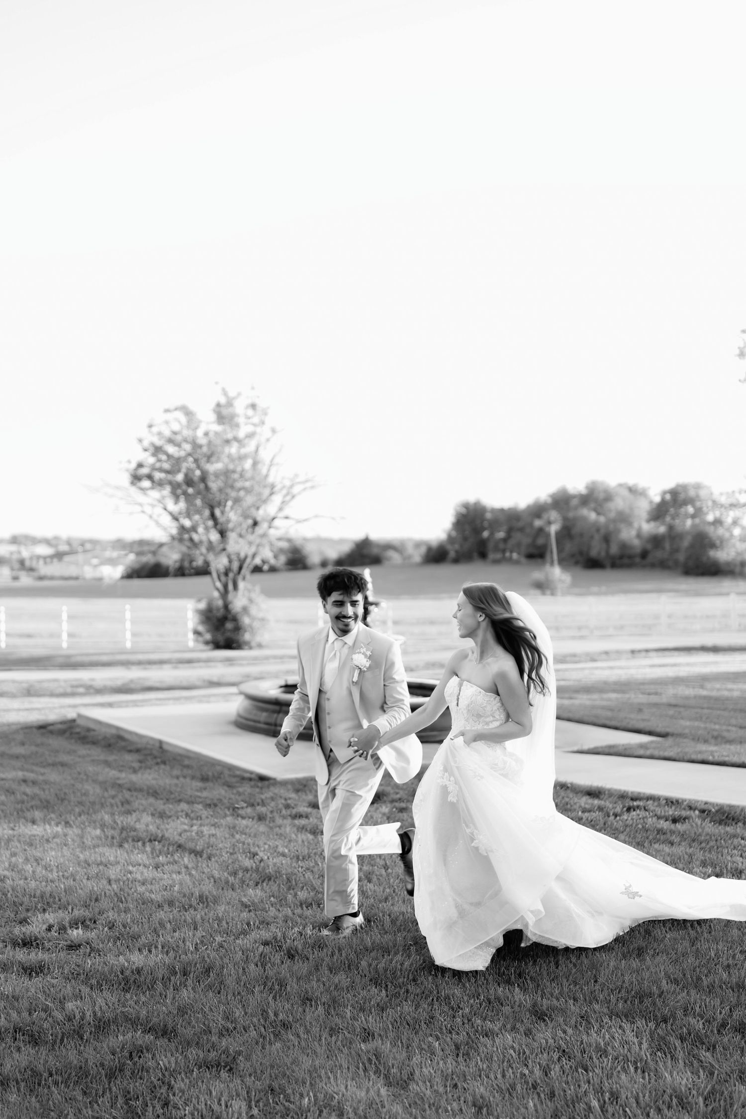 Black and white photo of a bride and groom holding hands, walking on grass at an outdoor wedding venue.