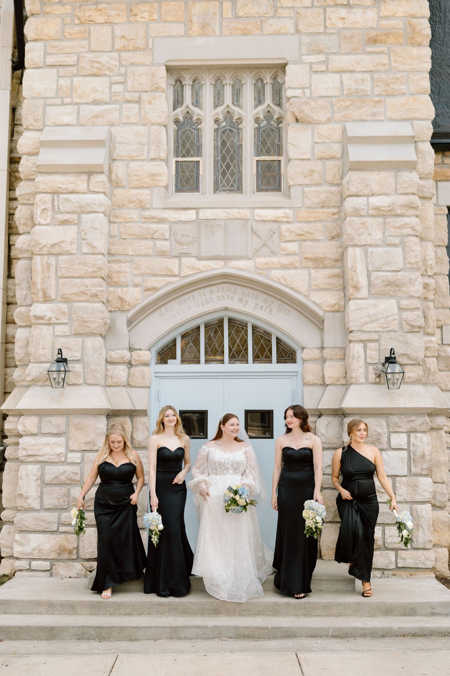 Bride in white gown with bridesmaids in black dresses walking in front of a stone church with a blue door.