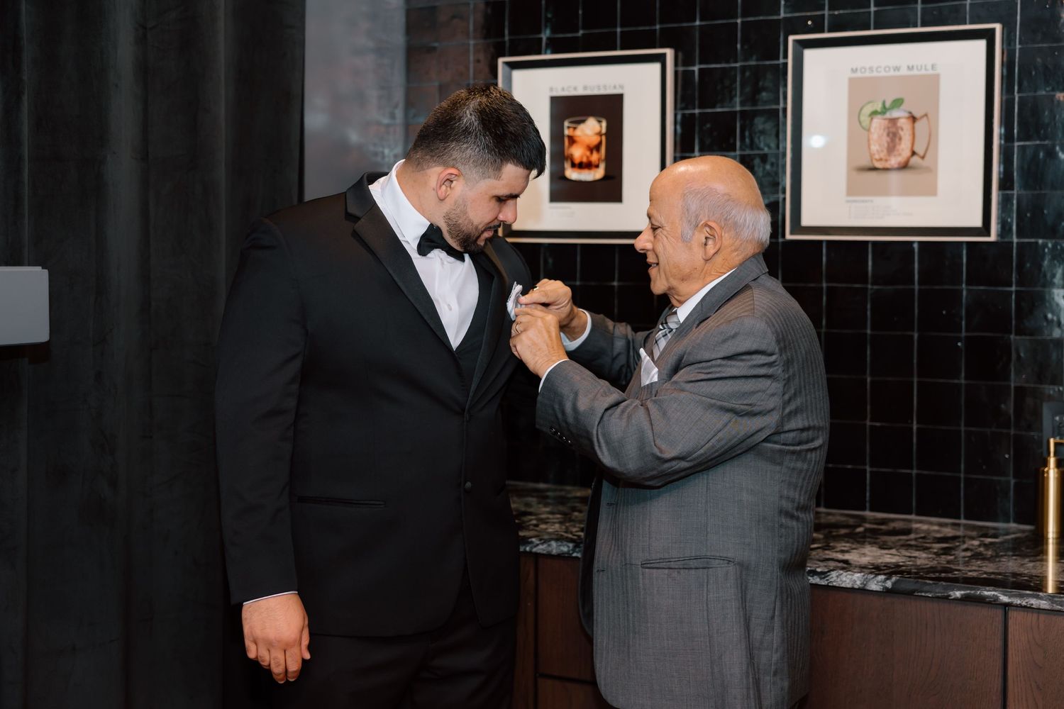 An elderly man adjusts a boutonniere on a groom in a black tuxedo at a wedding venue.