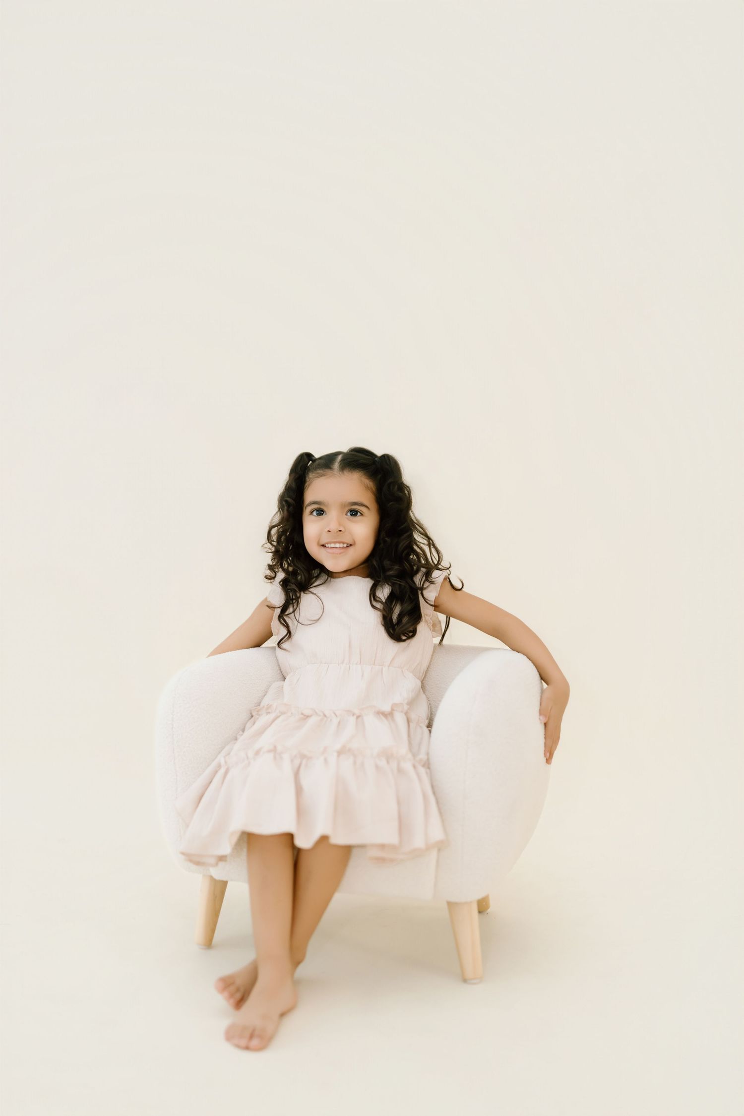 A child in a white dress sits on a white couch in a minimalist studio setting.