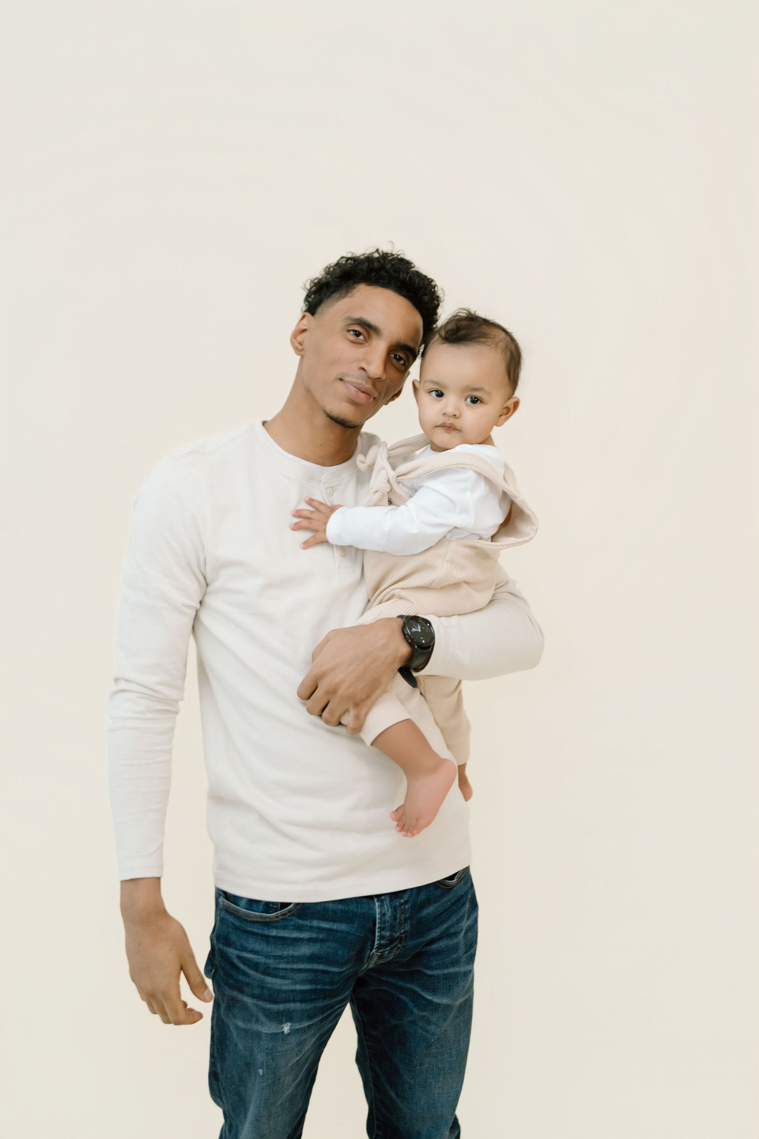 A person wearing a white long sleeve shirt and jeans holds a baby in white clothing against a light background.