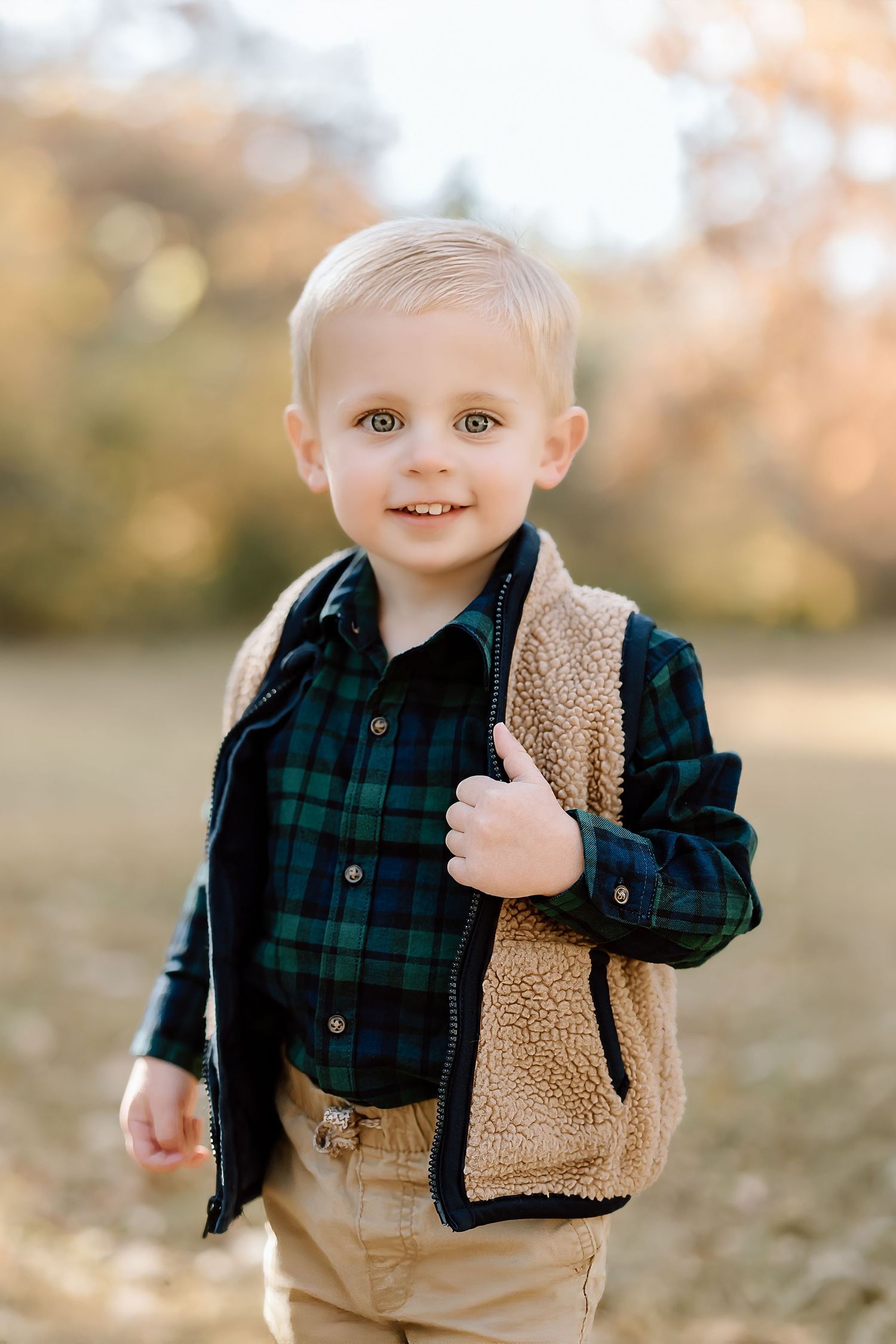 Child in plaid button-down and sherpa vest poses against blurred autumn background.