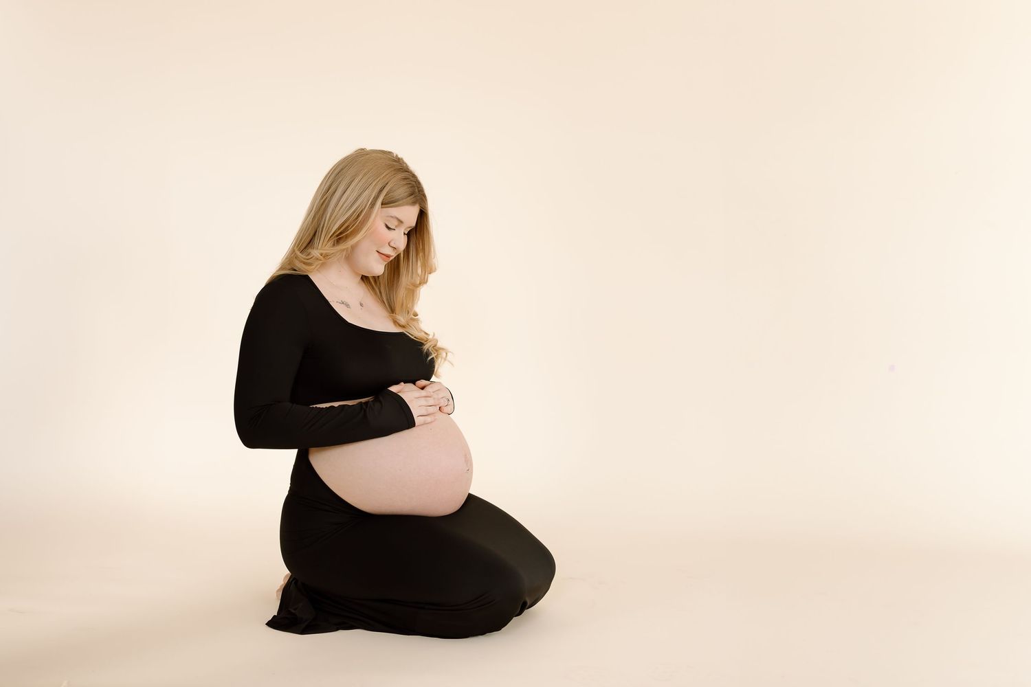 Gentle maternity pose with woman in black dress kneeling on cream colored studio floor.