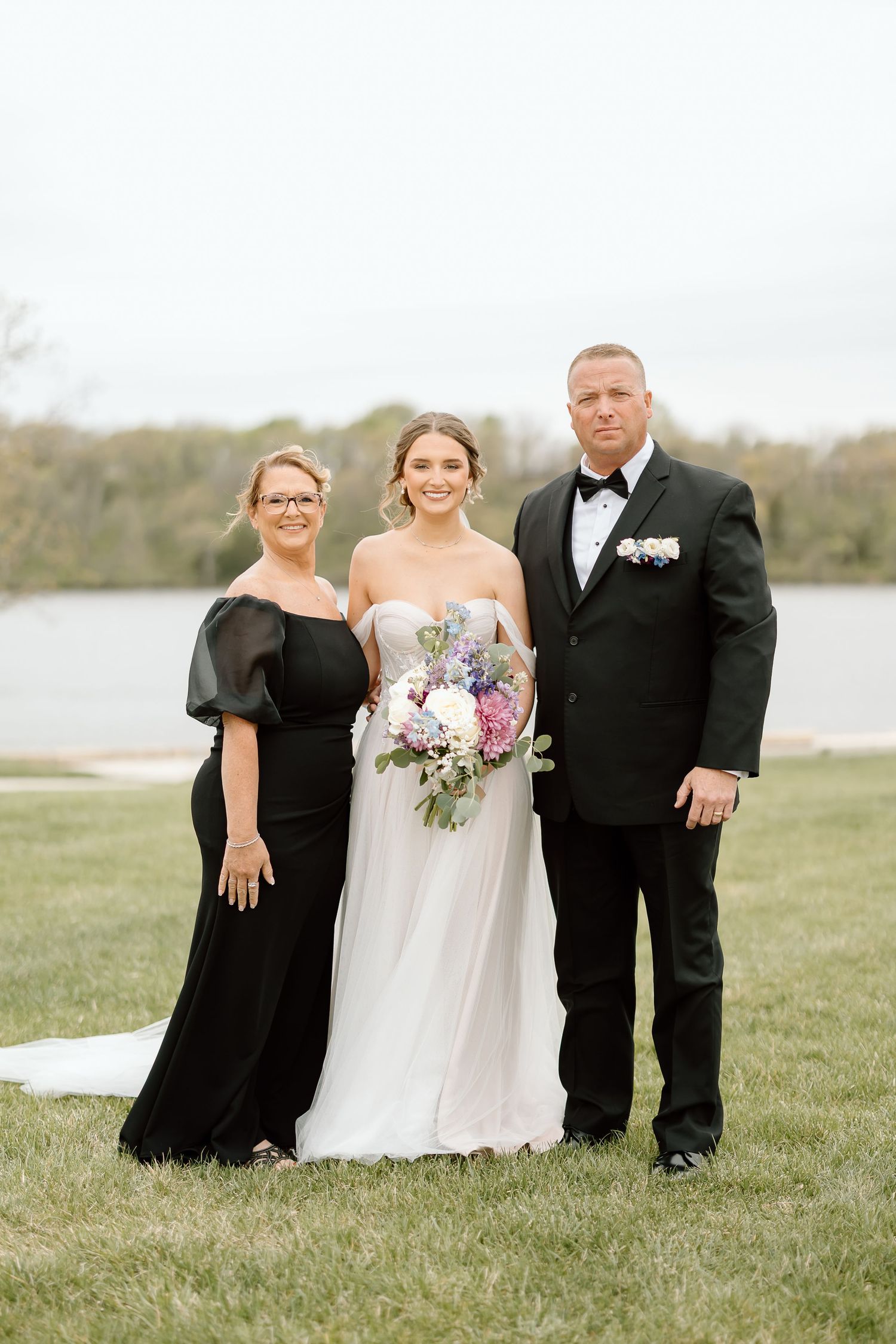 Formal wedding portrait with lake backdrop and purple floral bouquet.