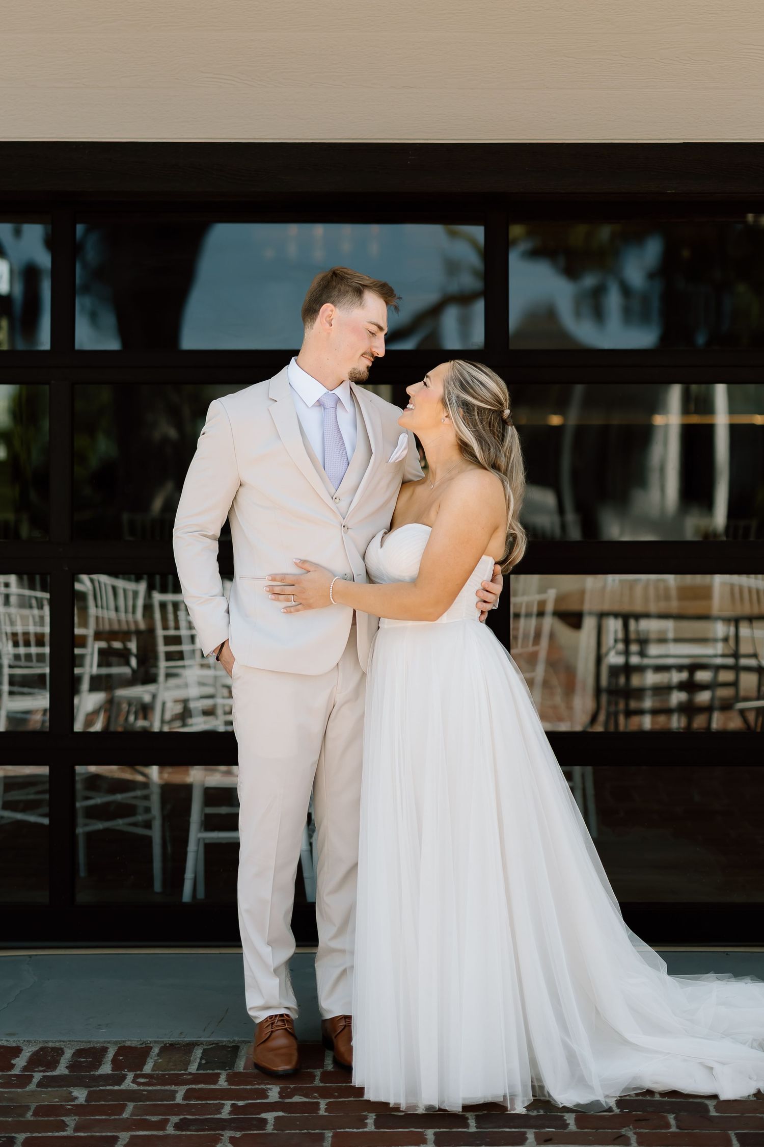 Couple embracing during first look outside venue with glass windows and brick walkway.