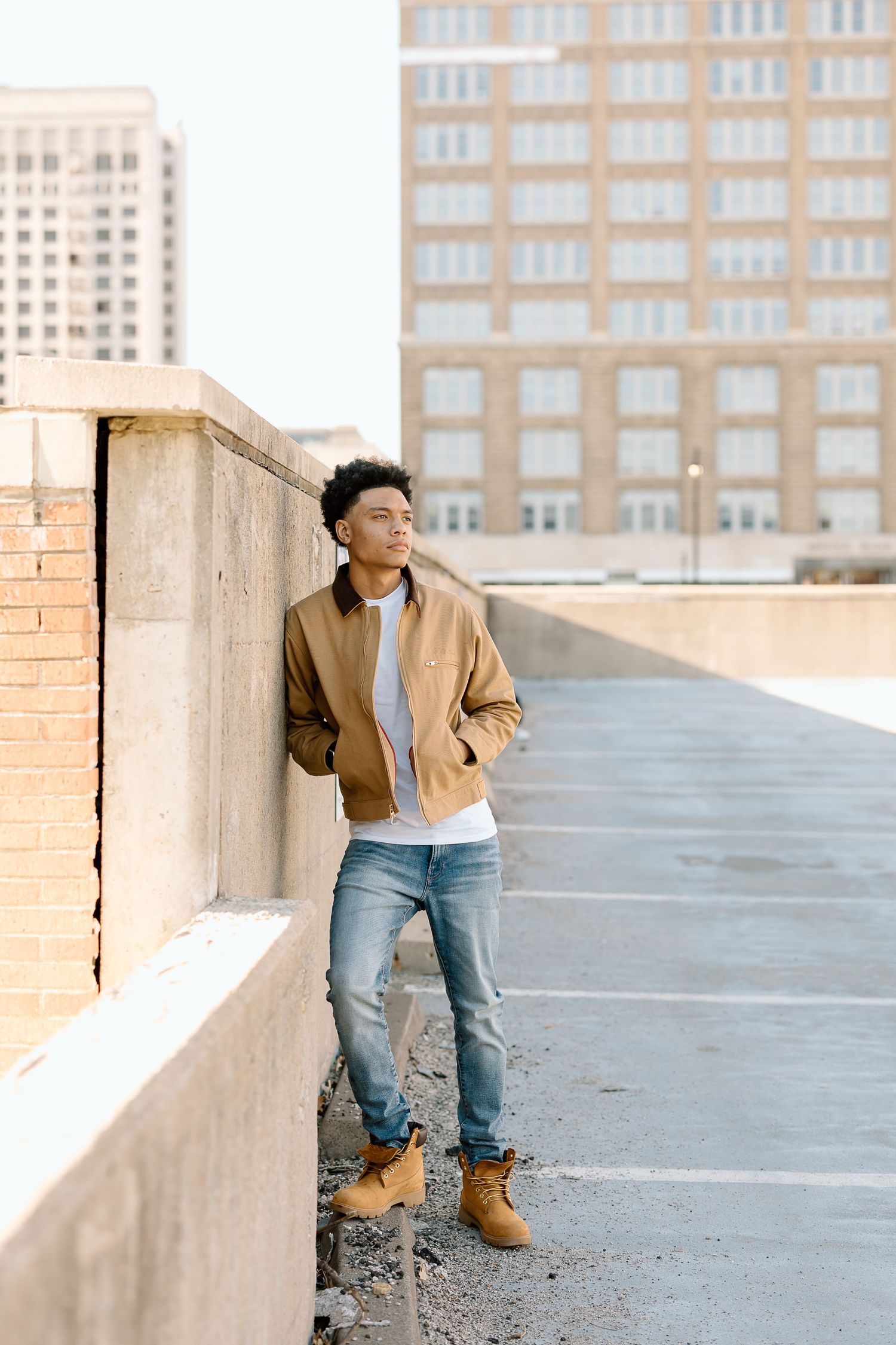 Urban fashion portrait showing casual style with tan jacket and jeans against city building backdrop.